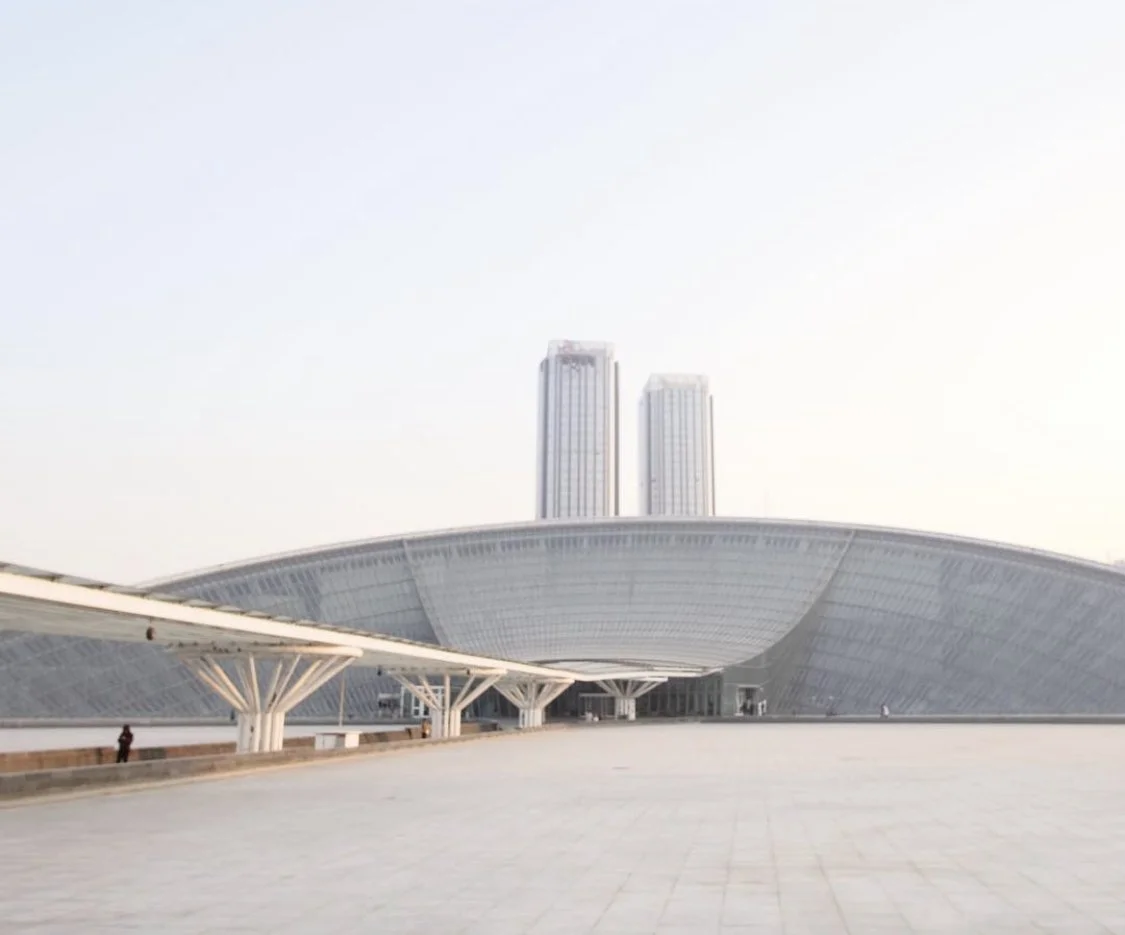 Modern architectural building with two tall towers and a curved roof structure in the foreground, under a clear sky.