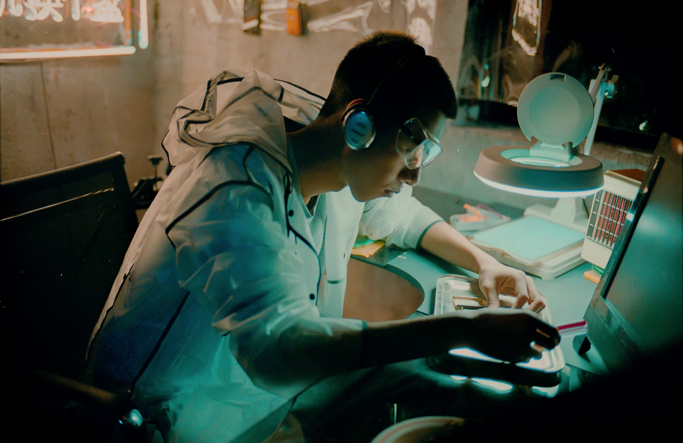 A young man wearing glasses, headphones, and a light-colored jacket working at his desk in a dimly lit room, focused on a small object in his hands, with a computer monitor, books, and a desk lamp nearby.