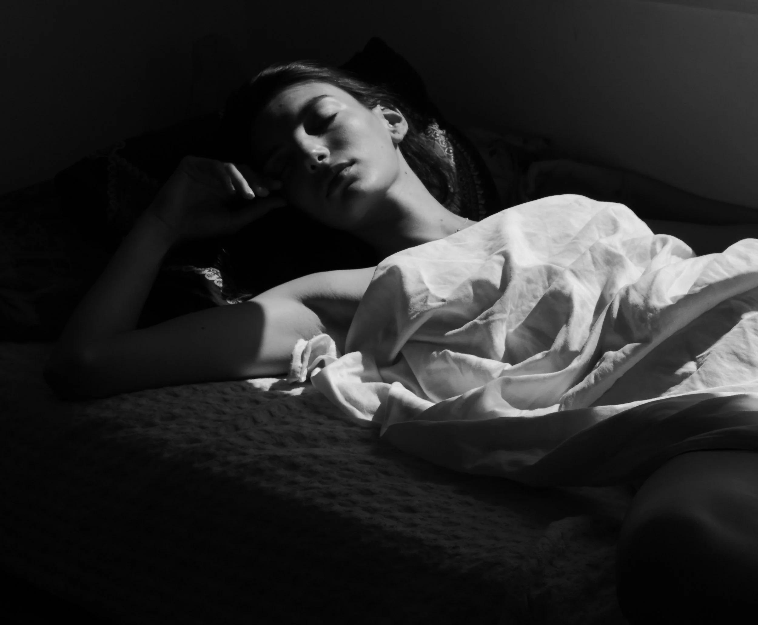 A woman sleeping on her side in a bed, covered partially with a blanket, in a dimly lit room