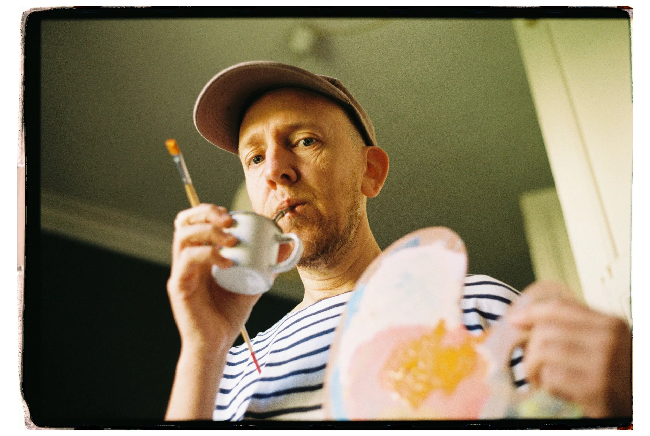 A man wearing a striped shirt and a baseball cap is holding a cup and bottle with a straw in his mouth, along with a colorful ceramic mug, in a room with a green ceiling and a white door.