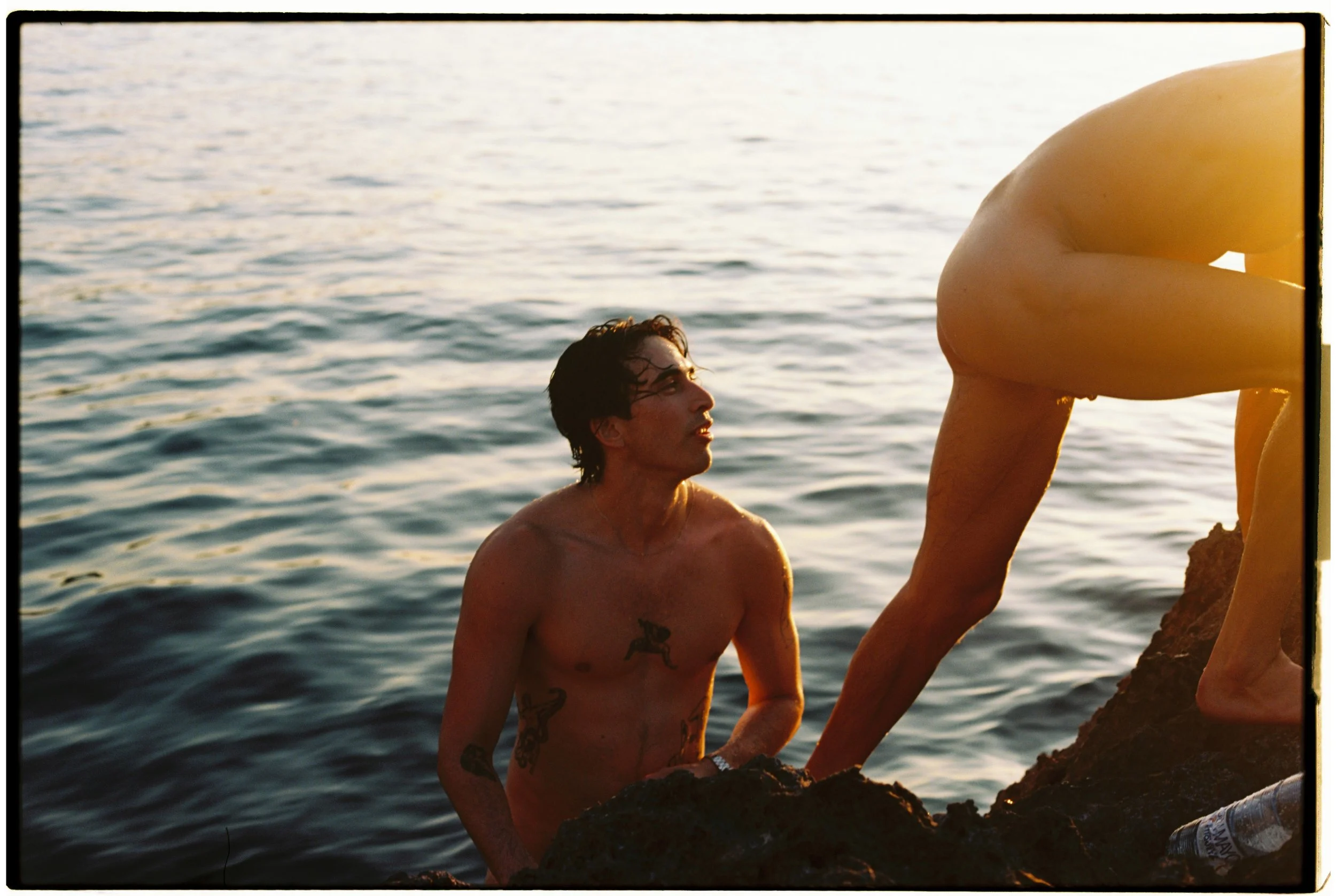 A shirtless man with tattoos on his chest and arms is in the water next to a rocky shoreline, looking at a person climbing out of the water onto the rocks during sunset.