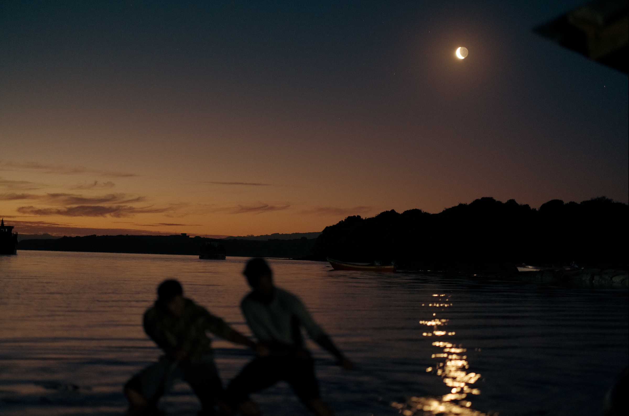 Two people fighting in a body of water during sunset or dusk, with boats and a hilly shoreline in the background, and the moon visible in the sky.