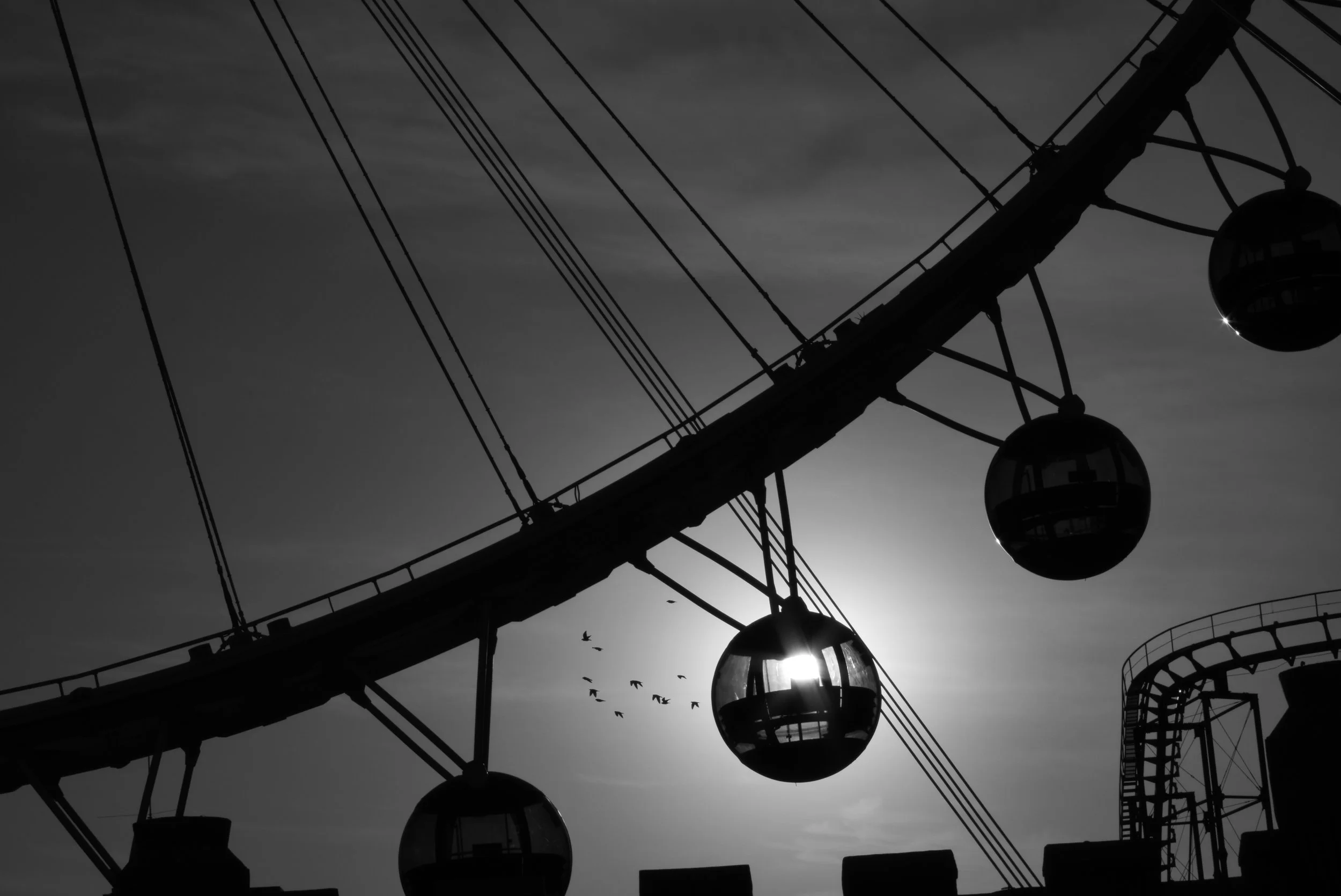 Silhouette of a Ferris wheel with spherical gondolas against the sun and sky, with birds flying nearby.