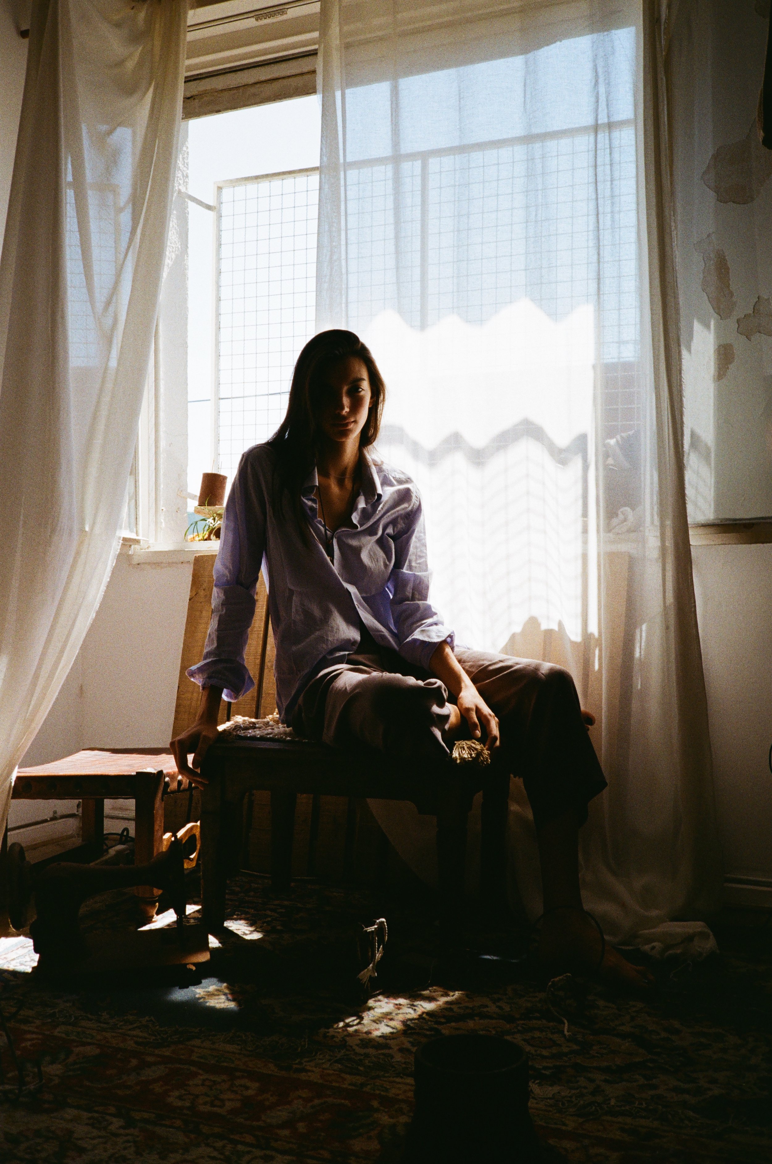 A woman sitting on a wooden chair in a room with sunlight streaming through a window with sheer curtains and a wire grid outside. The room has peeling paint on the wall and a patterned rug on the floor.
