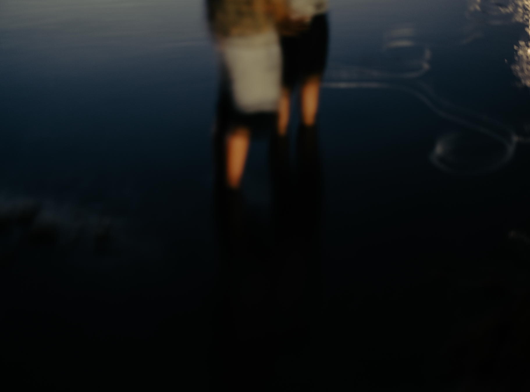 A person standing in shallow water at the beach, wearing a black swimsuit. The image is blurry and dark, with a reflection of the person visible in the water.