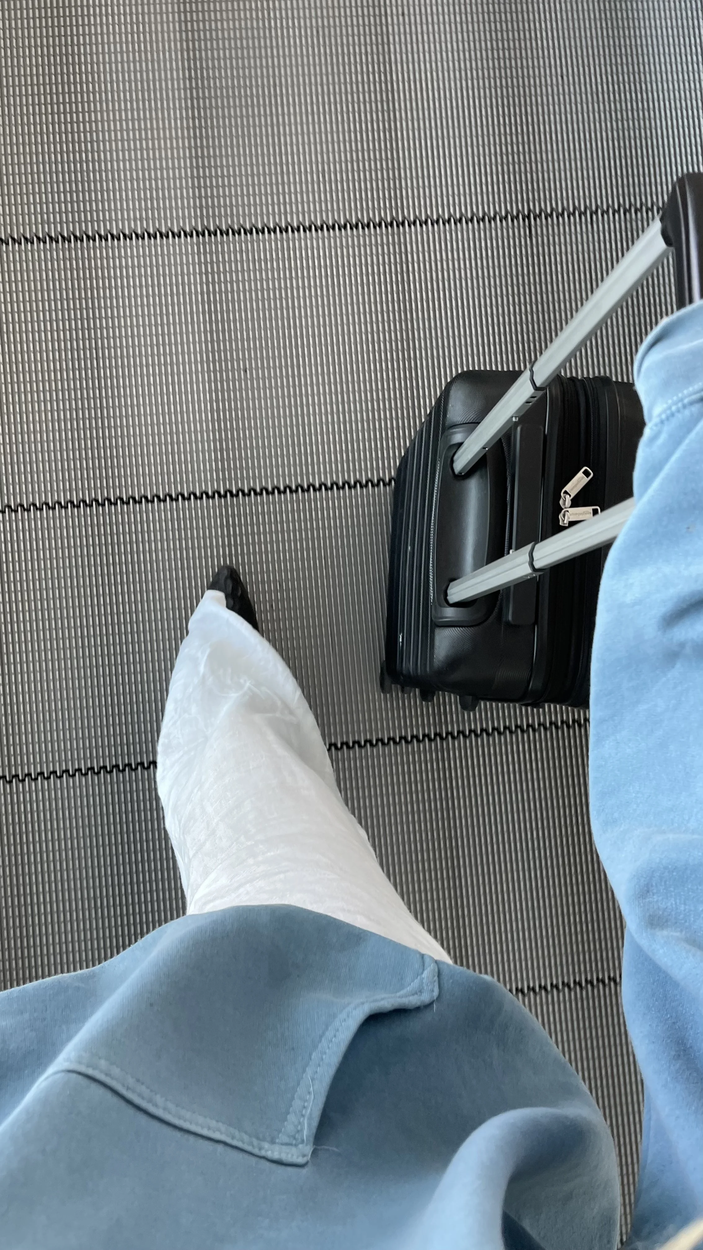 A person wearing light blue jeans and white pants with a light blue shirt is standing on an airport baggage claim carousel, holding a small black suitcase with an extendable handle.
