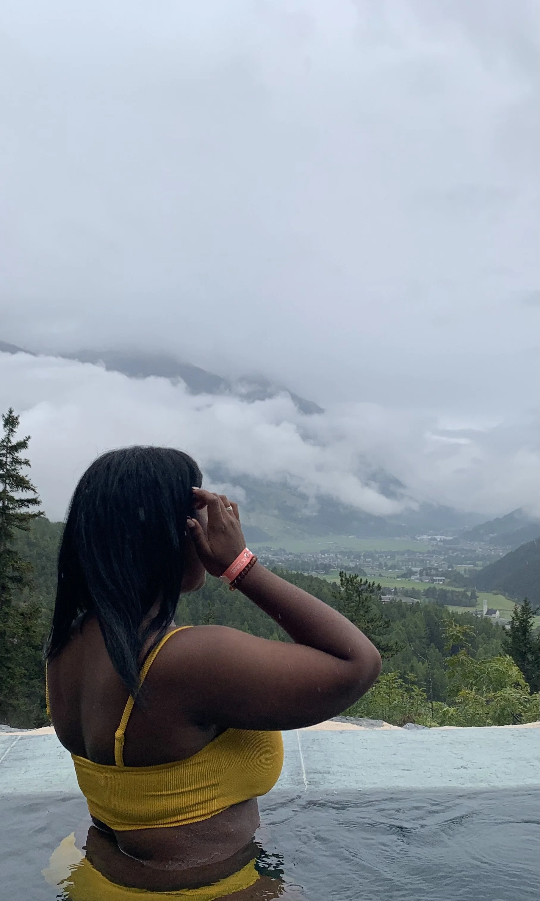 Woman in yellow swimsuit standing in a hot spring or pool, overlooking a cloudy mountain landscape with green valley below.