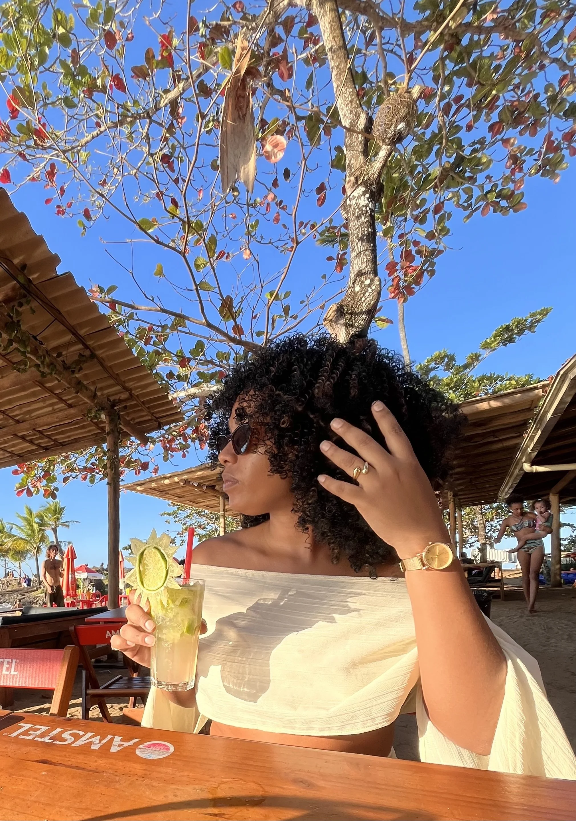 A woman with curly black hair and sunglasses sitting at an outdoor beach bar, holding a cocktail with lime slices, under a blue sky and orange, palm, and red-leafed trees in the background.