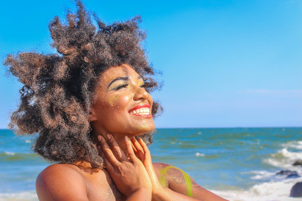 Smiling woman with curly hair on a beach, wearing colorful makeup and glitter, with the ocean in the background.