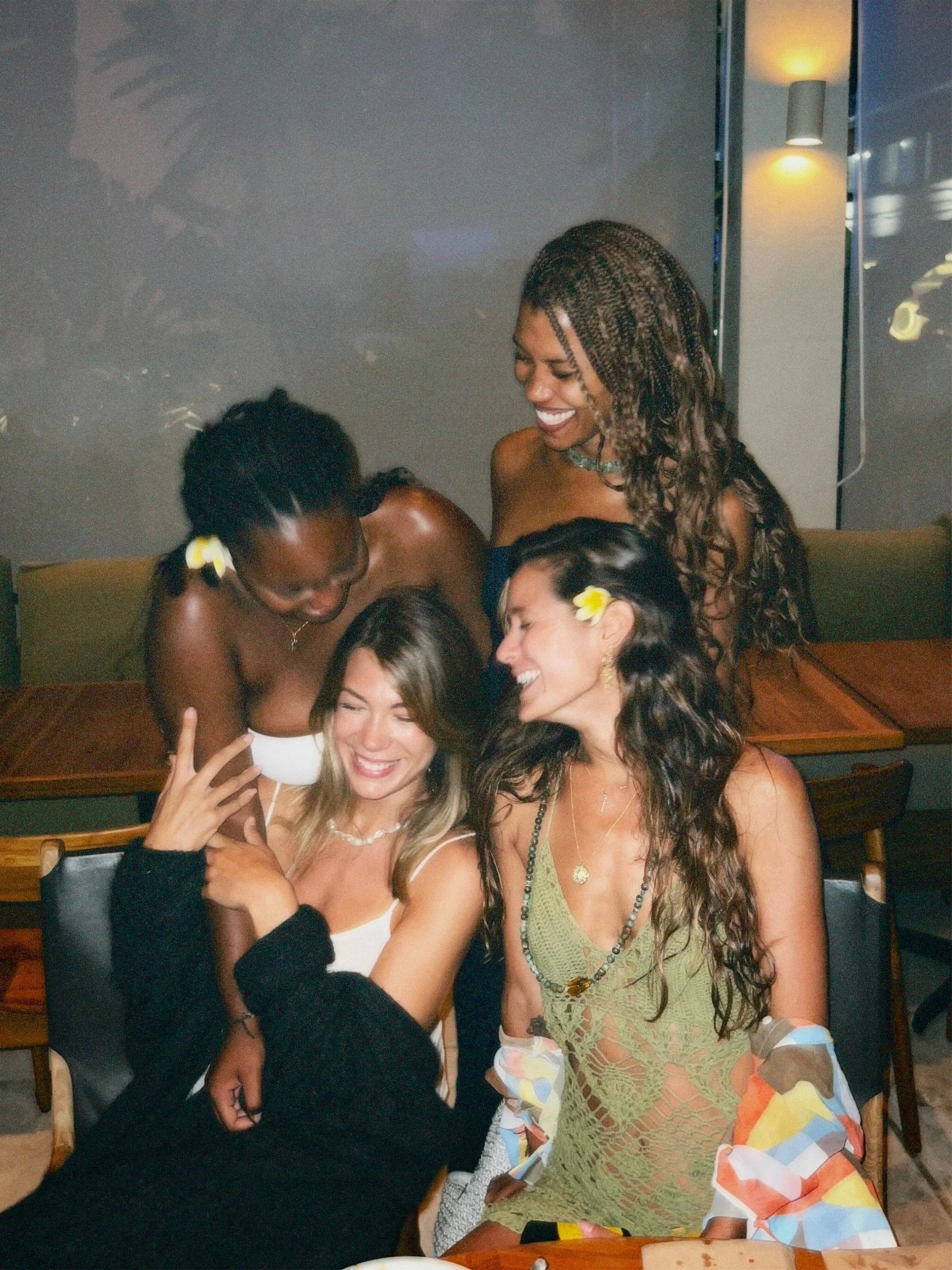 Five women enjoying a moment together at a restaurant, smiling and laughing, with some wearing flower accessories in their hair.
