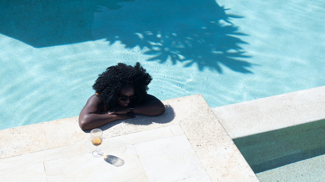 Woman with curly hair wearing sunglasses relaxing by a swimming pool with a glass of wine on the ledge.