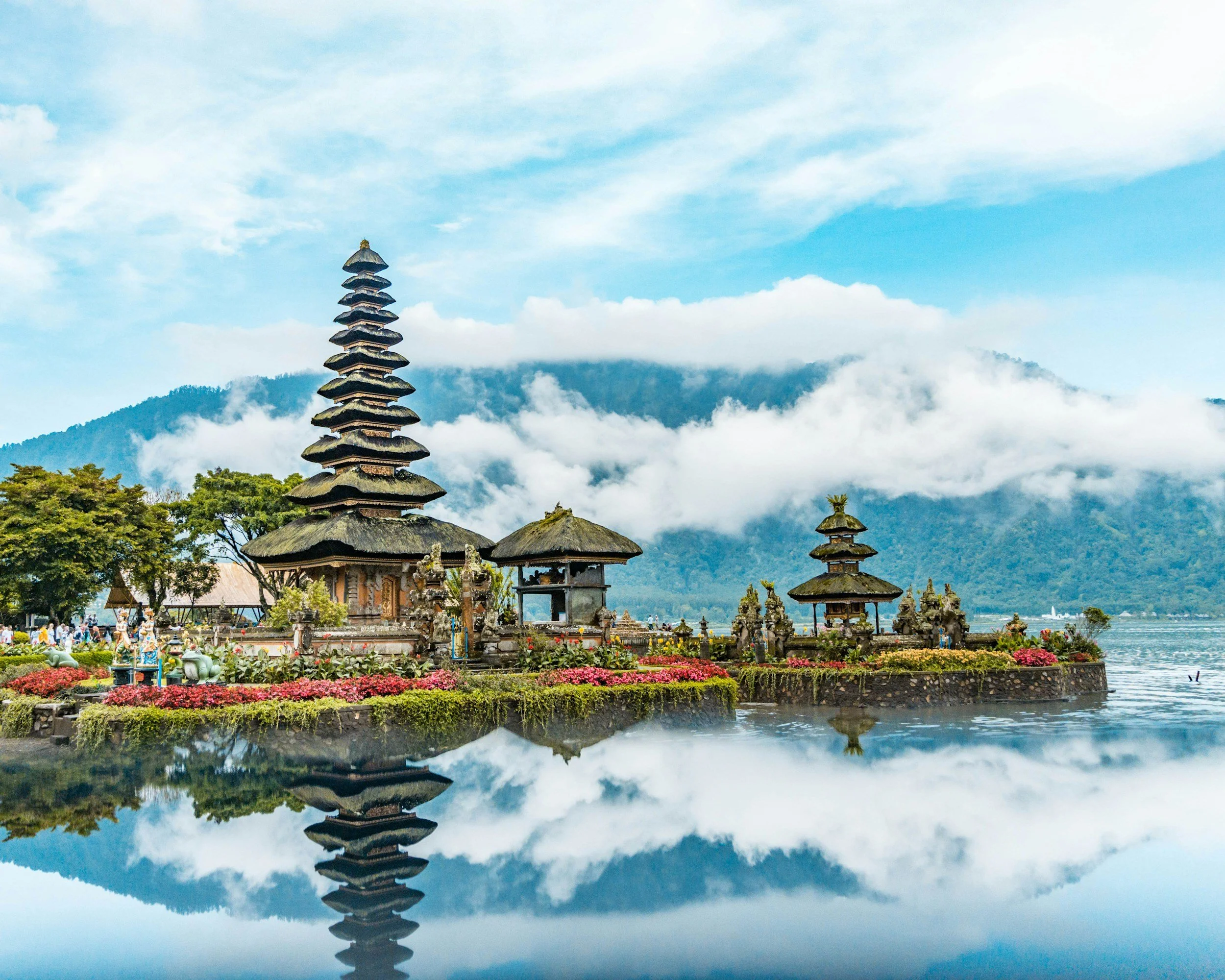 Traditional Balinese temple structures with tiered roofs near a body of water, surrounded by colorful flowers, with mountains and clouds in the background.