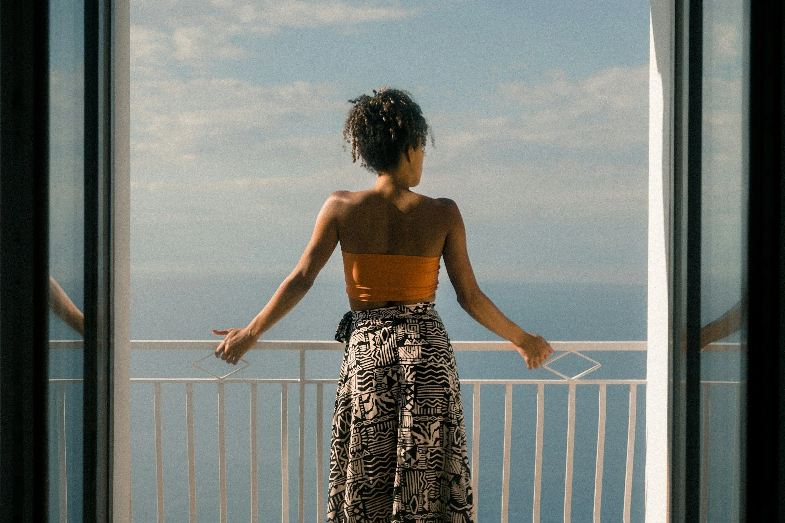 A woman with curly hair stands on a balcony, facing the ocean, holding a hanger in her right hand, wearing a strapless orange top and patterned wide-leg pants.