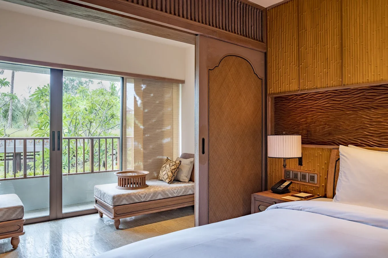 A hotel room with sliding glass doors leading to a balcony with greenery outside, featuring a daybed with patterned cushions, and a bed with a wooden headboard, a nightstand with a lamp and telephone.