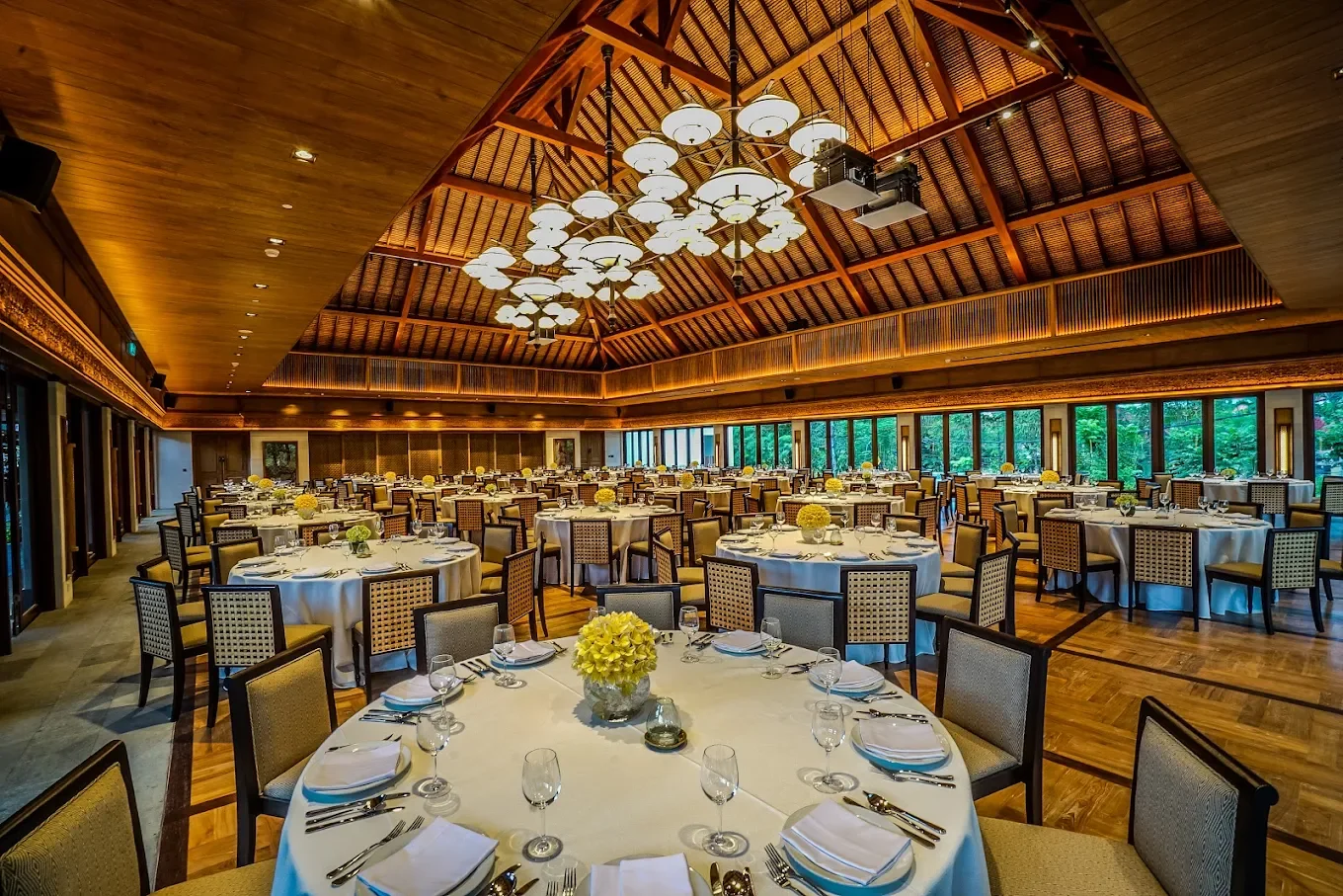 Elegant banquet hall with multiple round tables set for a formal event, featuring white tablecloths, floral centerpieces, and neatly arranged place settings. The high wooden ceiling has modern pendant lighting, and large windows line the walls, providing a view of lush greenery outside.