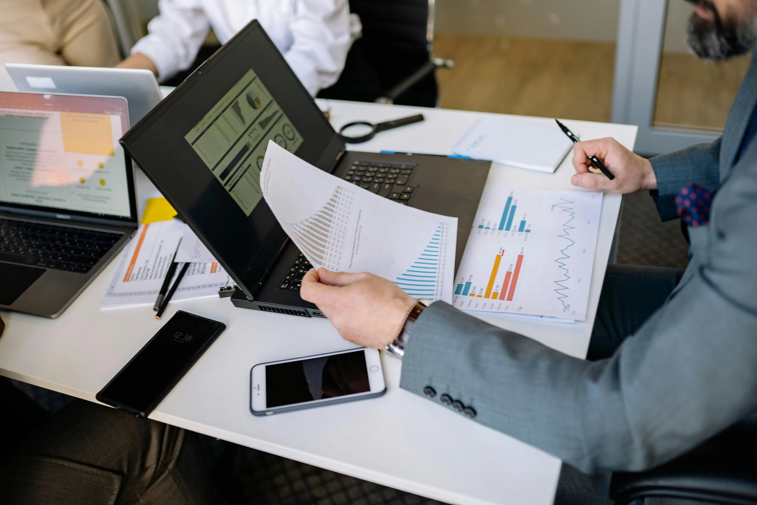 Business meeting with documents and laptops on a table, showing charts and graphs, with one person holding a paper and another person using a pen.