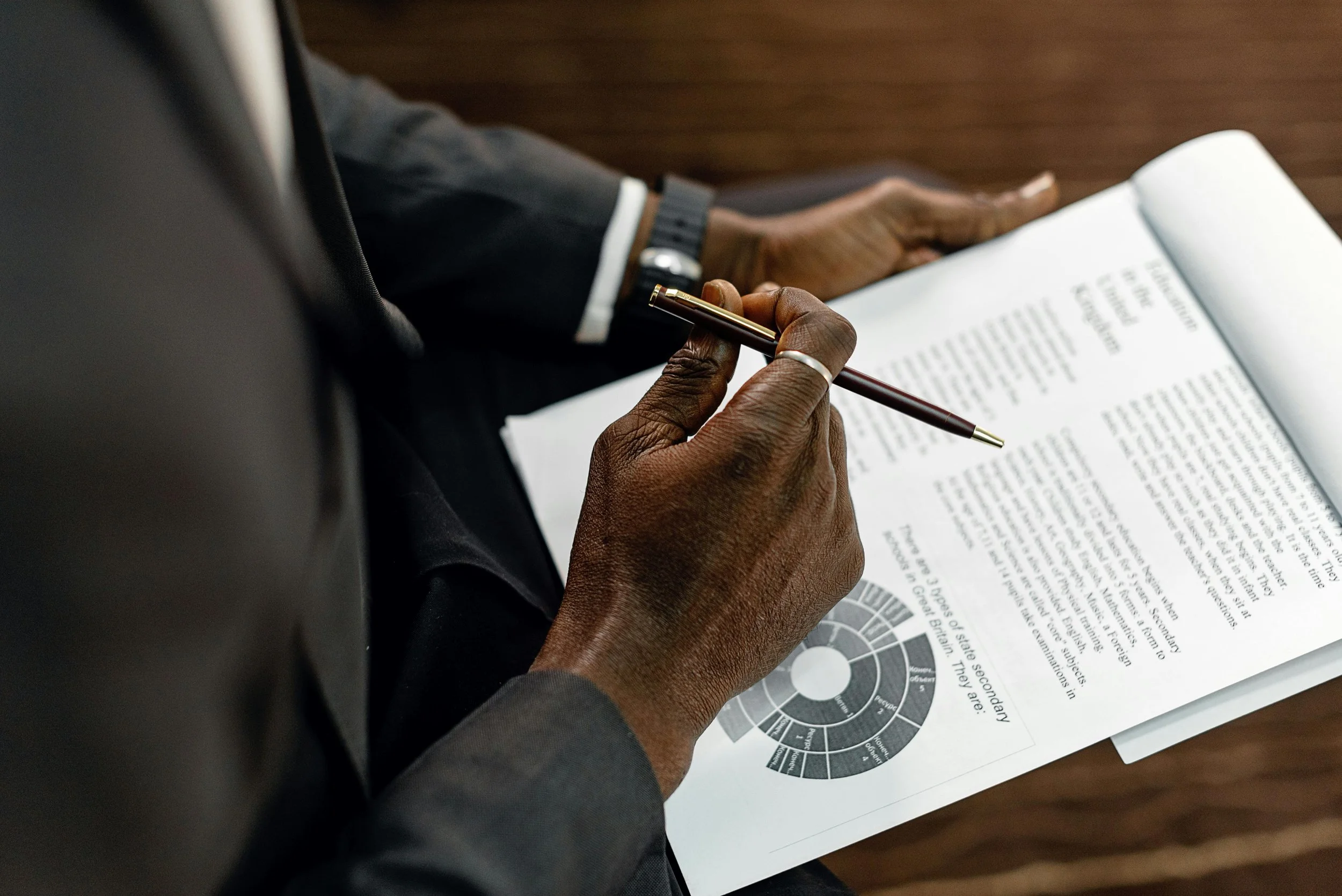A person dressed in a business suit is holding a pen and reviewing a printed document with text and a circular diagram or chart.