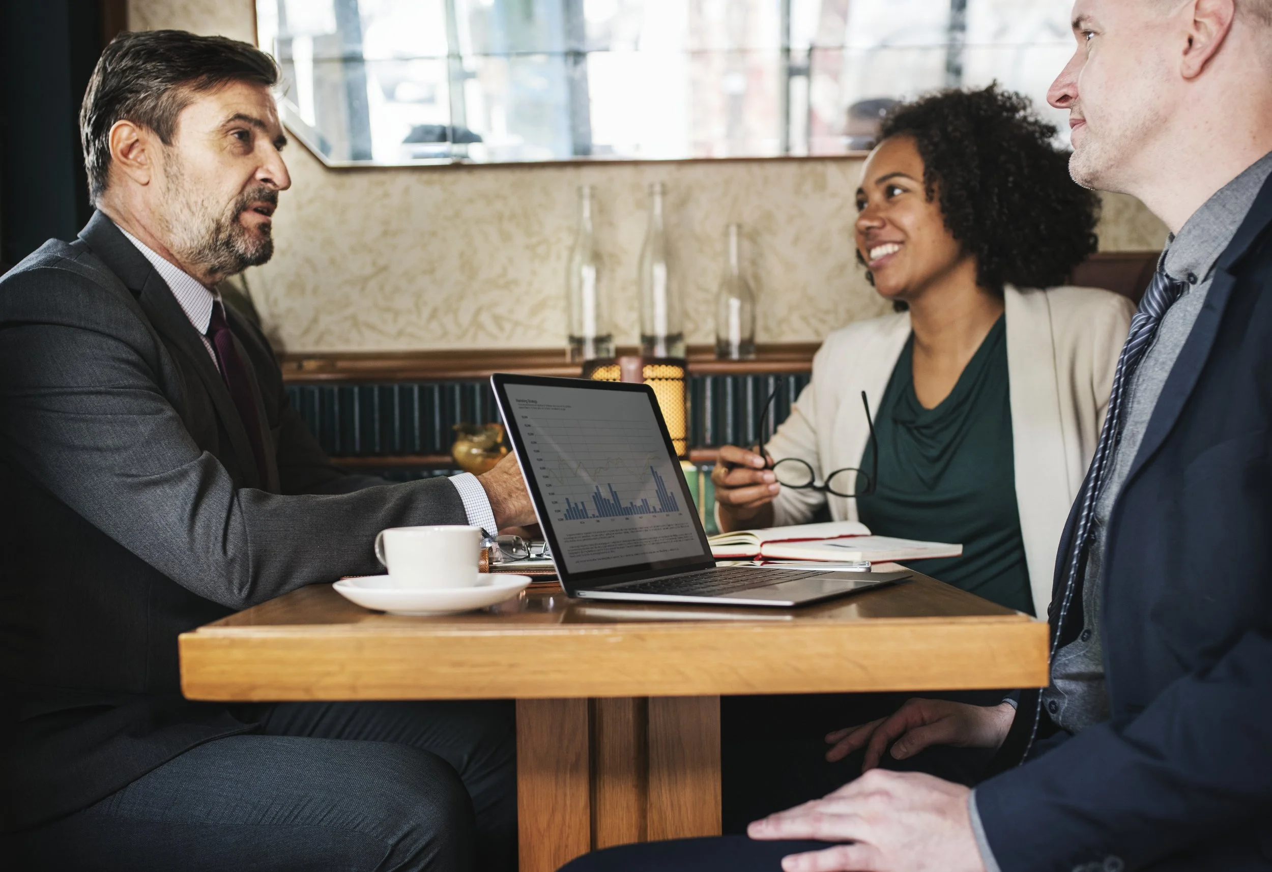 Four professionals having a meeting at a restaurant, with a man in a suit speaking and three others listening, a laptop displaying graphs and charts on the table.