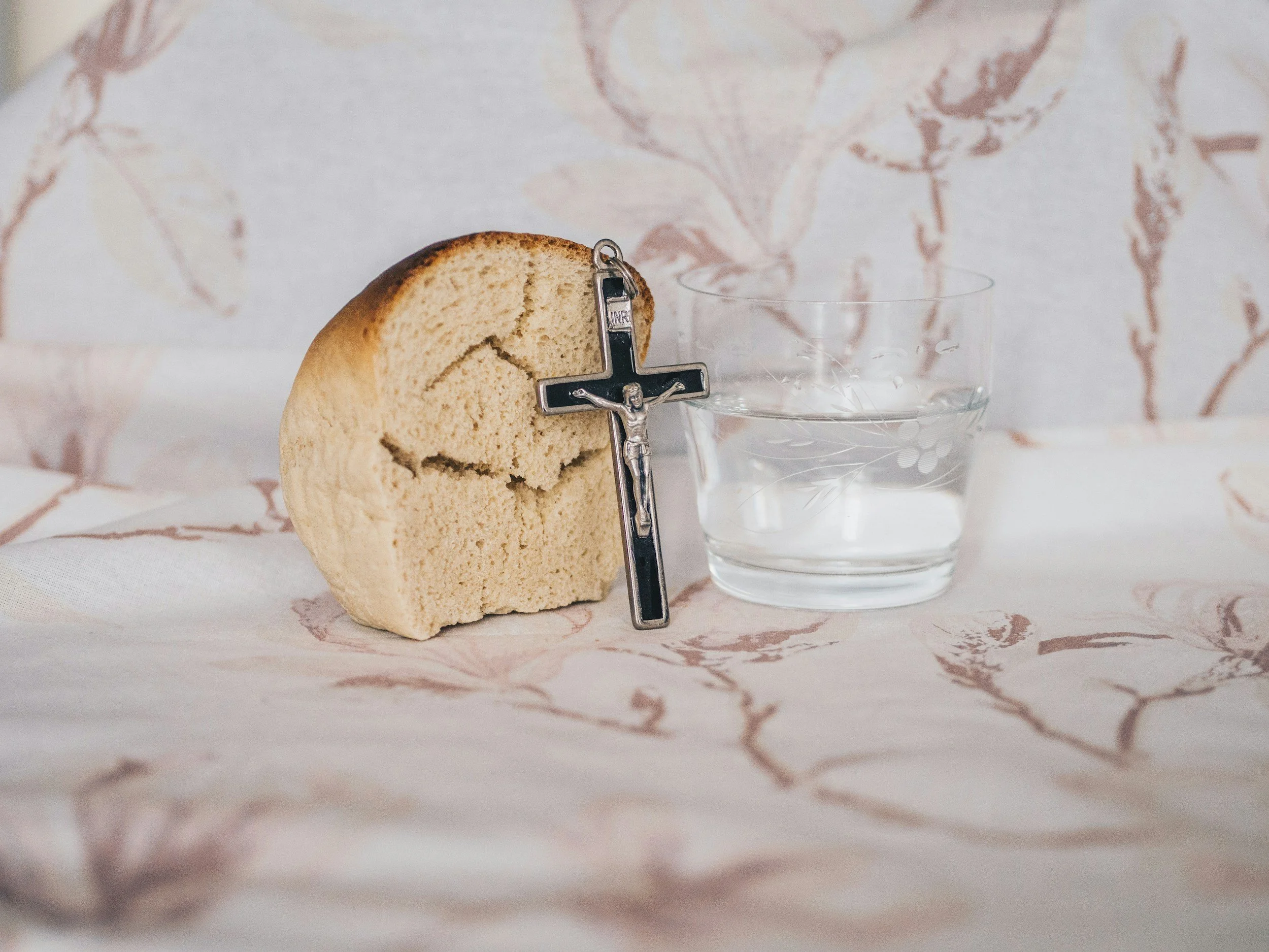 A round piece of bread with a crucifix leaning against it, and a glass of water next to the bread, set on a surface with a floral pattern background.