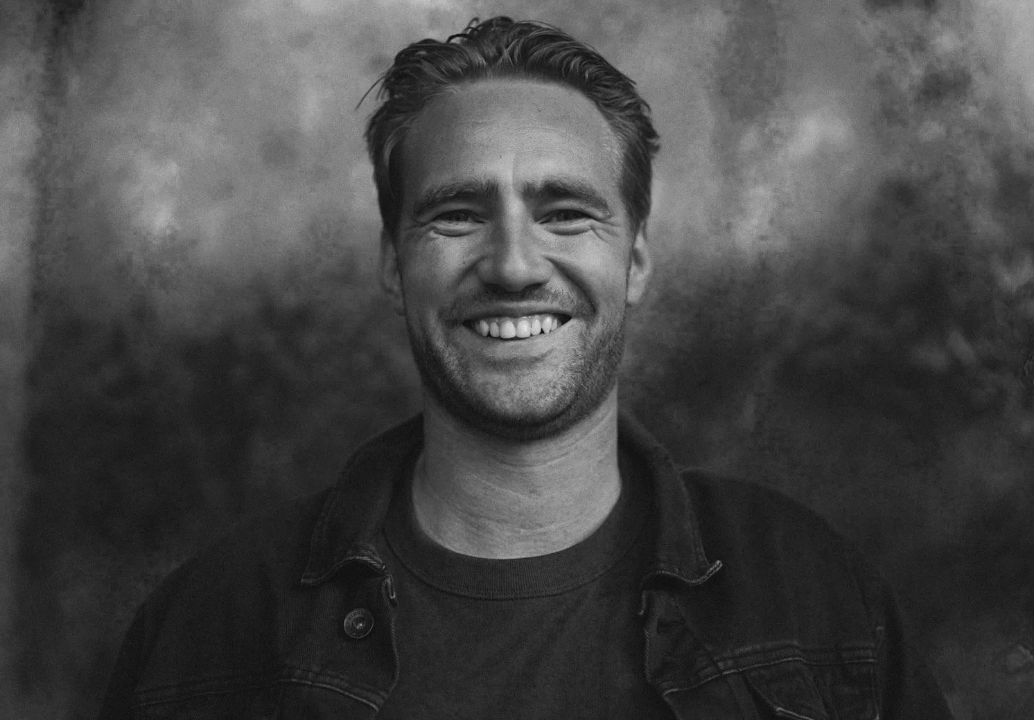 Black and white portrait of a man smiling, wearing a denim jacket over a t-shirt, with a textured background.