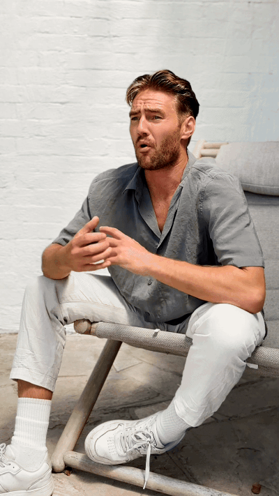 A man sitting on a modern lounge chair against a white brick wall, wearing a gray shirt, white pants, and white sneakers, talking and gesturing with his hands.