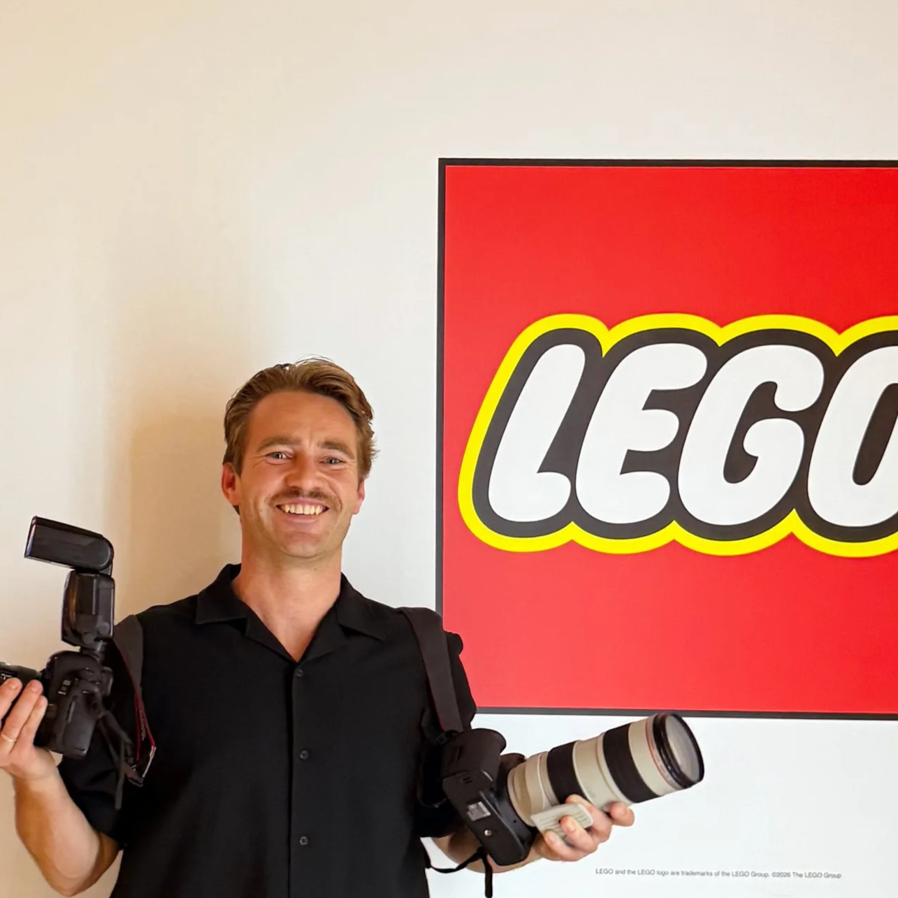 A man holding a camera with a telephoto lens, standing next to a large LEGO logo sign on a white wall.