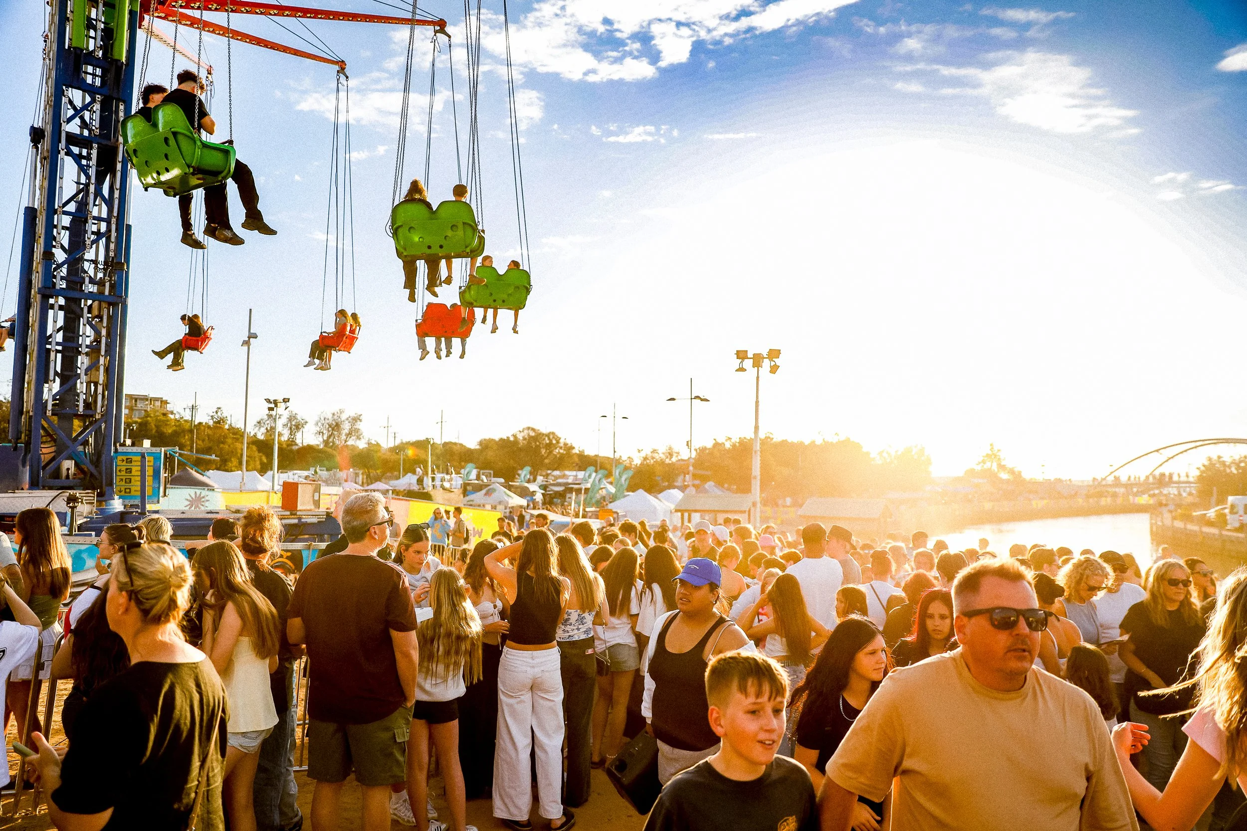 A crowded outdoor fair at sunset with a crowd of people, some wearing sunglasses, near a children's swing ride with kids in bright-colored seats. The sky is partly cloudy and the sunlight creates a warm glow.