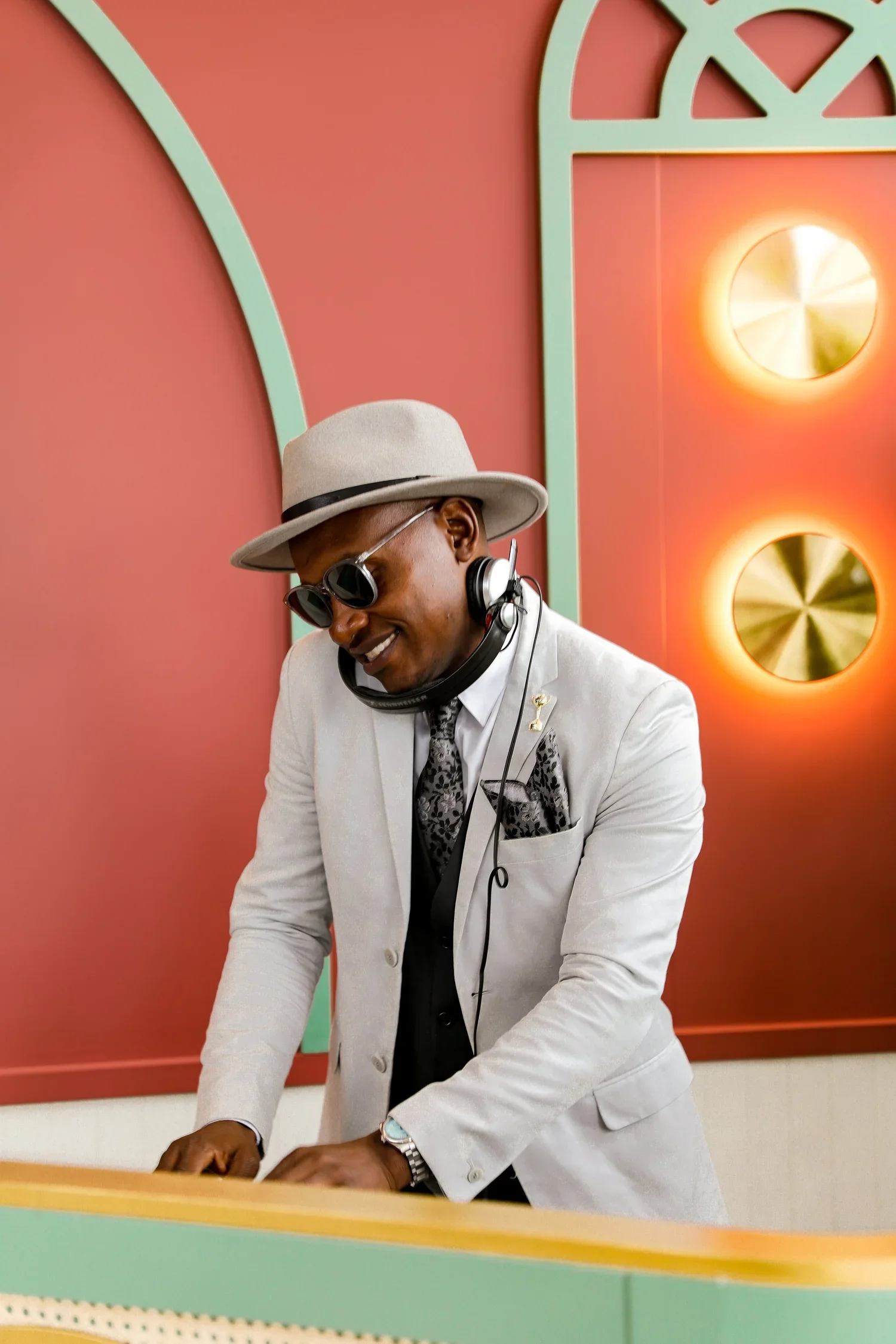 A man dressed in a light gray suit, black vest, printed pocket square, and white hat, playing a keyboard with headphones and sunglasses, smiling in a colorful room with pink walls and decorative gold-colored circular wall accents.