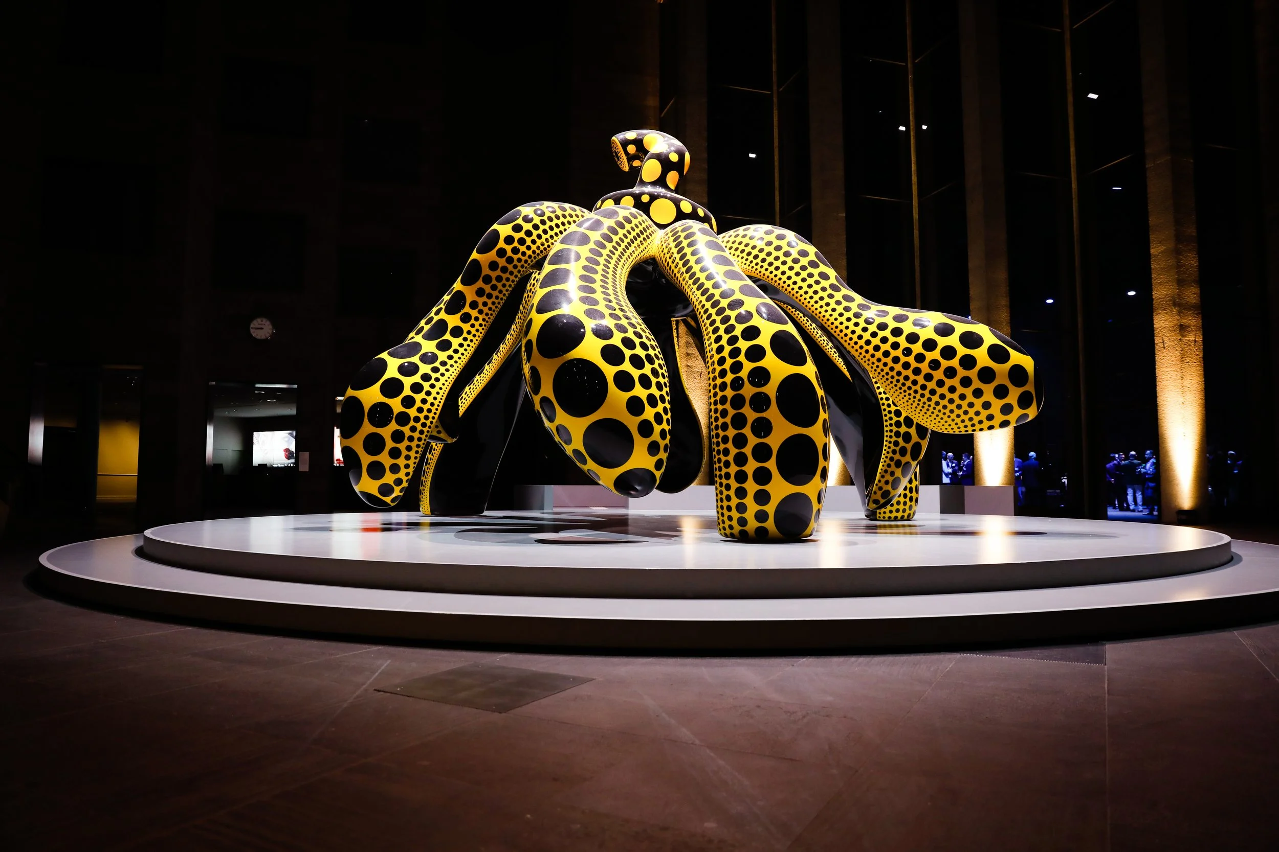 A large, abstract sculpture resembling an octopus or squid with long, rounded tentacles covered in a yellow and black polka dot pattern, displayed on a circular white platform in a dark, modern indoor space.