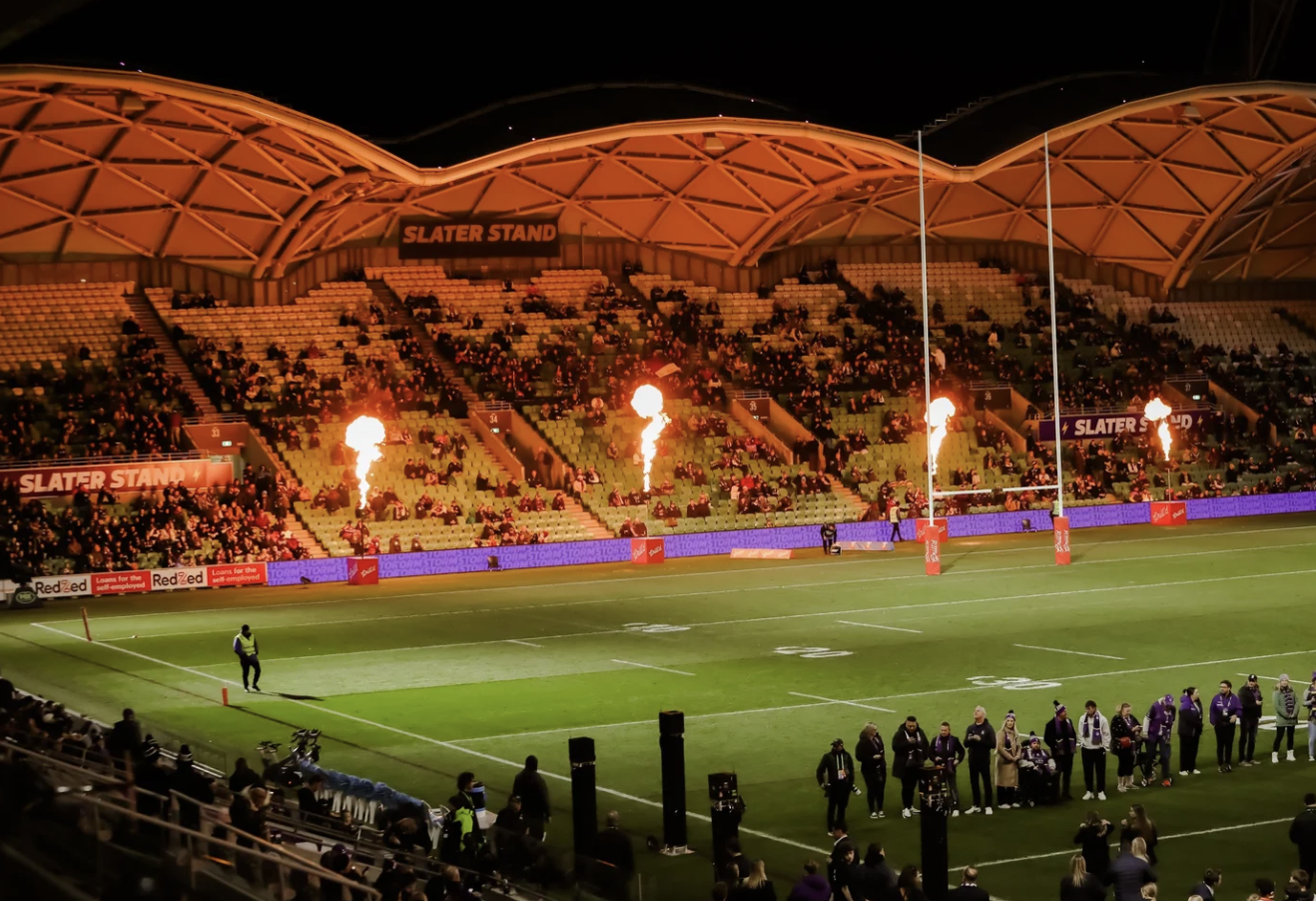 Nighttime sports stadium with spectators, flaming torches on the stands, and a well-lit field.