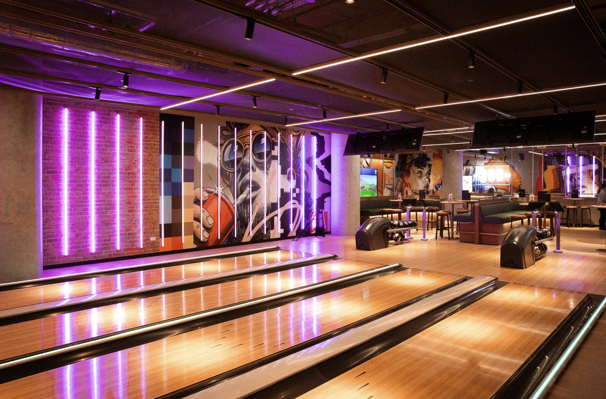 A modern bowling alley with four lanes, neon purple lighting, and colorful urban artwork on the walls.