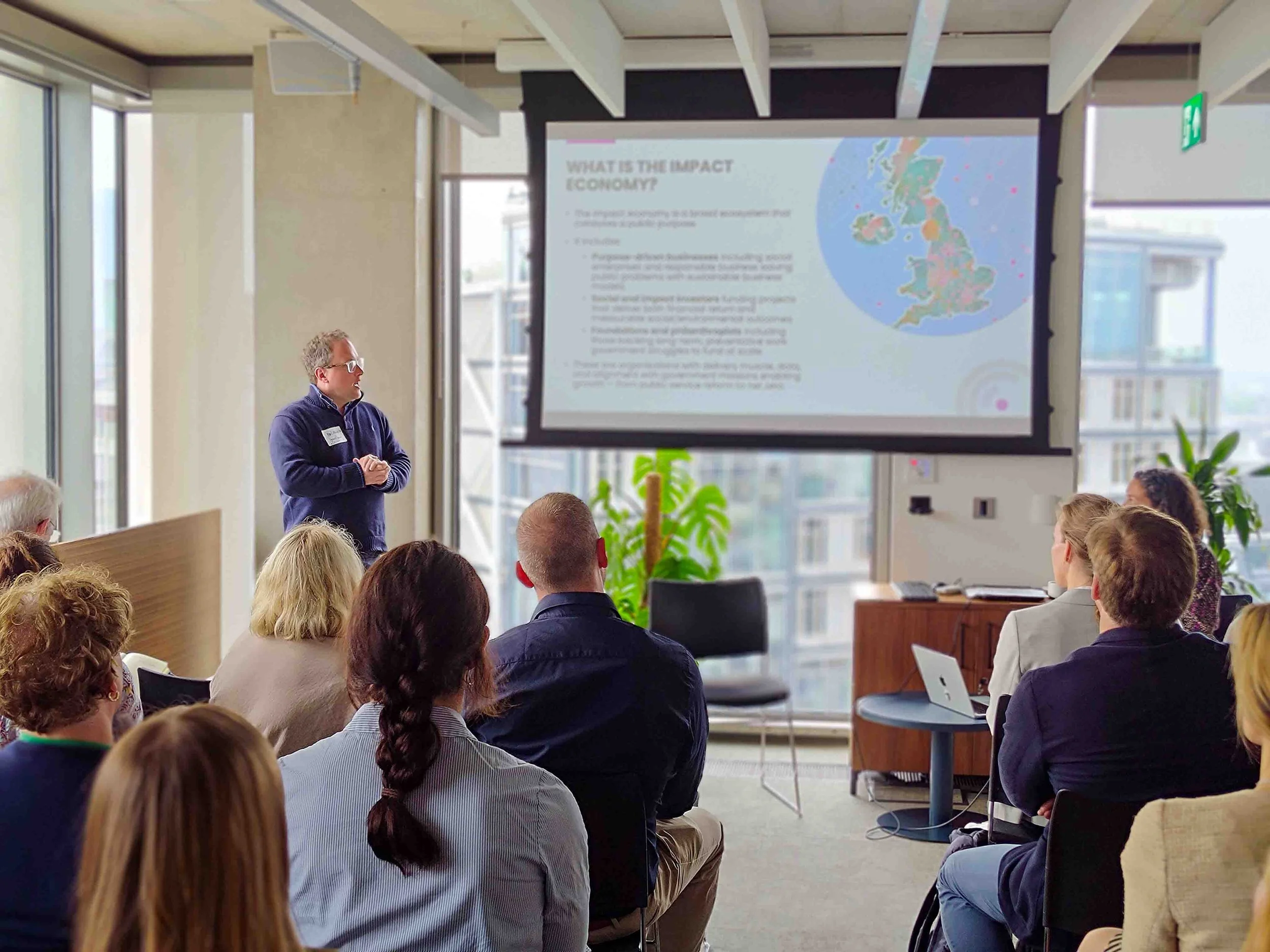 Man giving a presentation to an audience in a bright conference room with large windows and a projector screen displaying a slide titled 'What is the impact on the economy?'