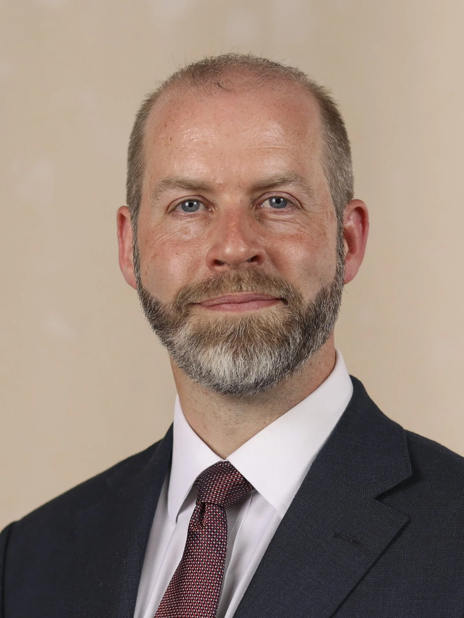 A professional headshot of a middle-aged man with a trimmed beard and short hair, wearing a dark suit, white shirt, and patterned tie, standing against a beige background.