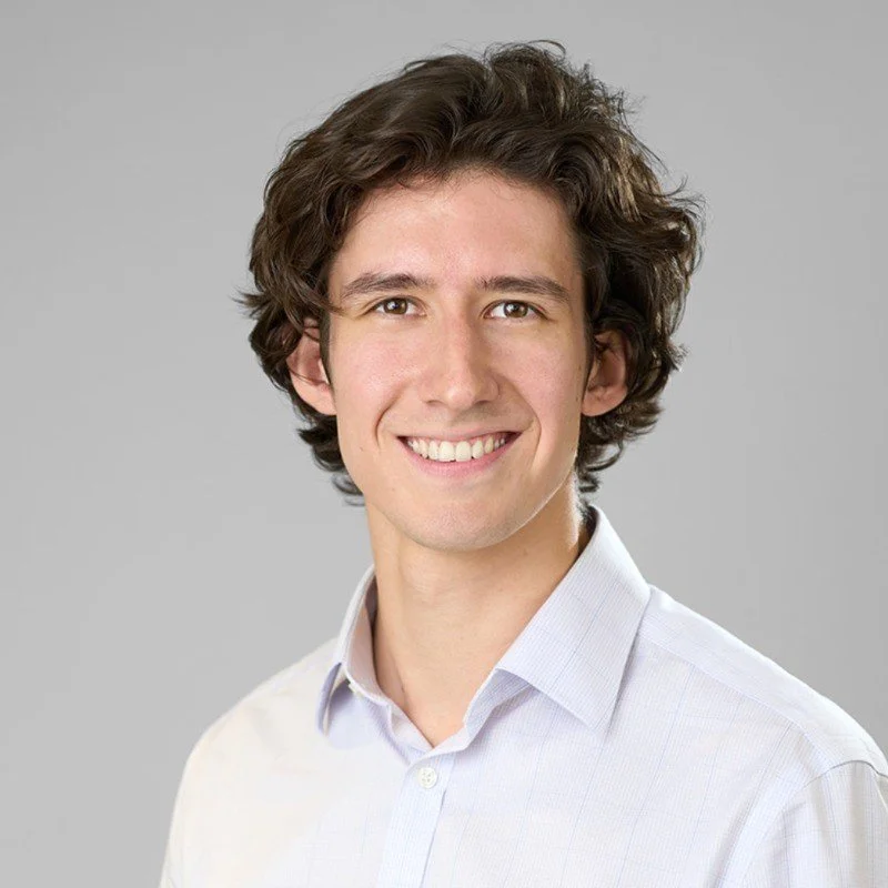 A young man with curly brown hair, smiling, wearing a light-colored dress shirt against a plain gray background.