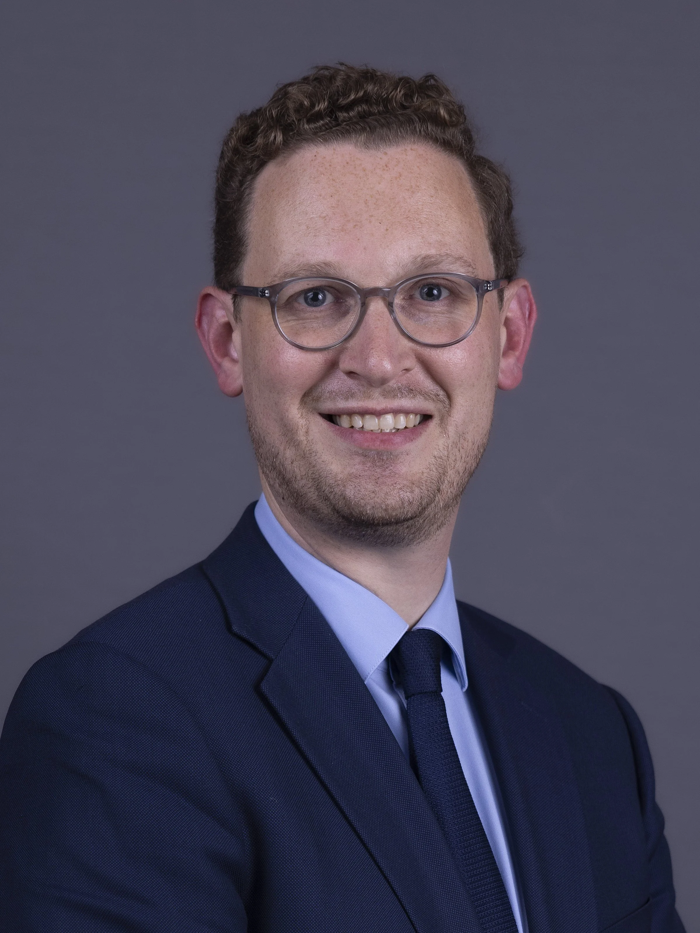 A professional headshot of a man with short, curly brown hair, wearing glasses, a blue dress shirt, a dark tie, and a dark blazer, smiling against a gray background.