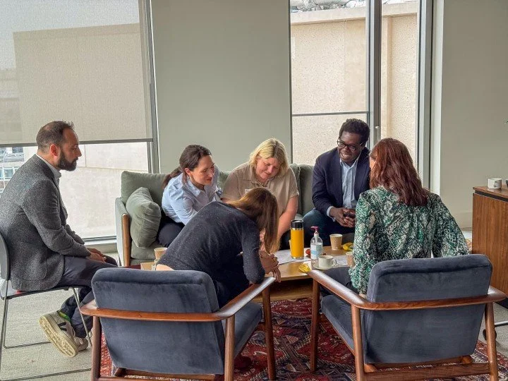 A group of six diverse people engaged in a discussion around a coffee table in a modern office meeting room with large windows.