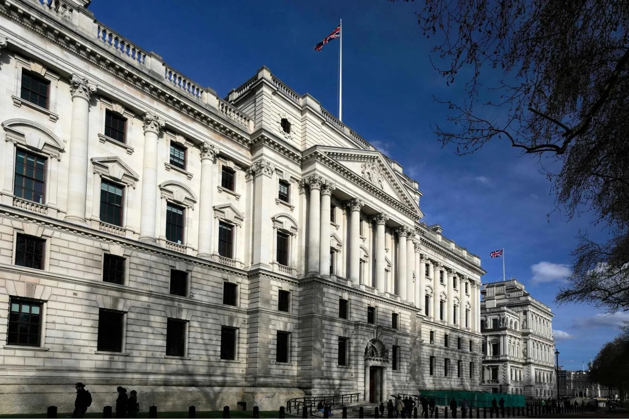White historic government building with columns, windows, and Union Jack flags on the roof, under a blue sky with some clouds and trees.