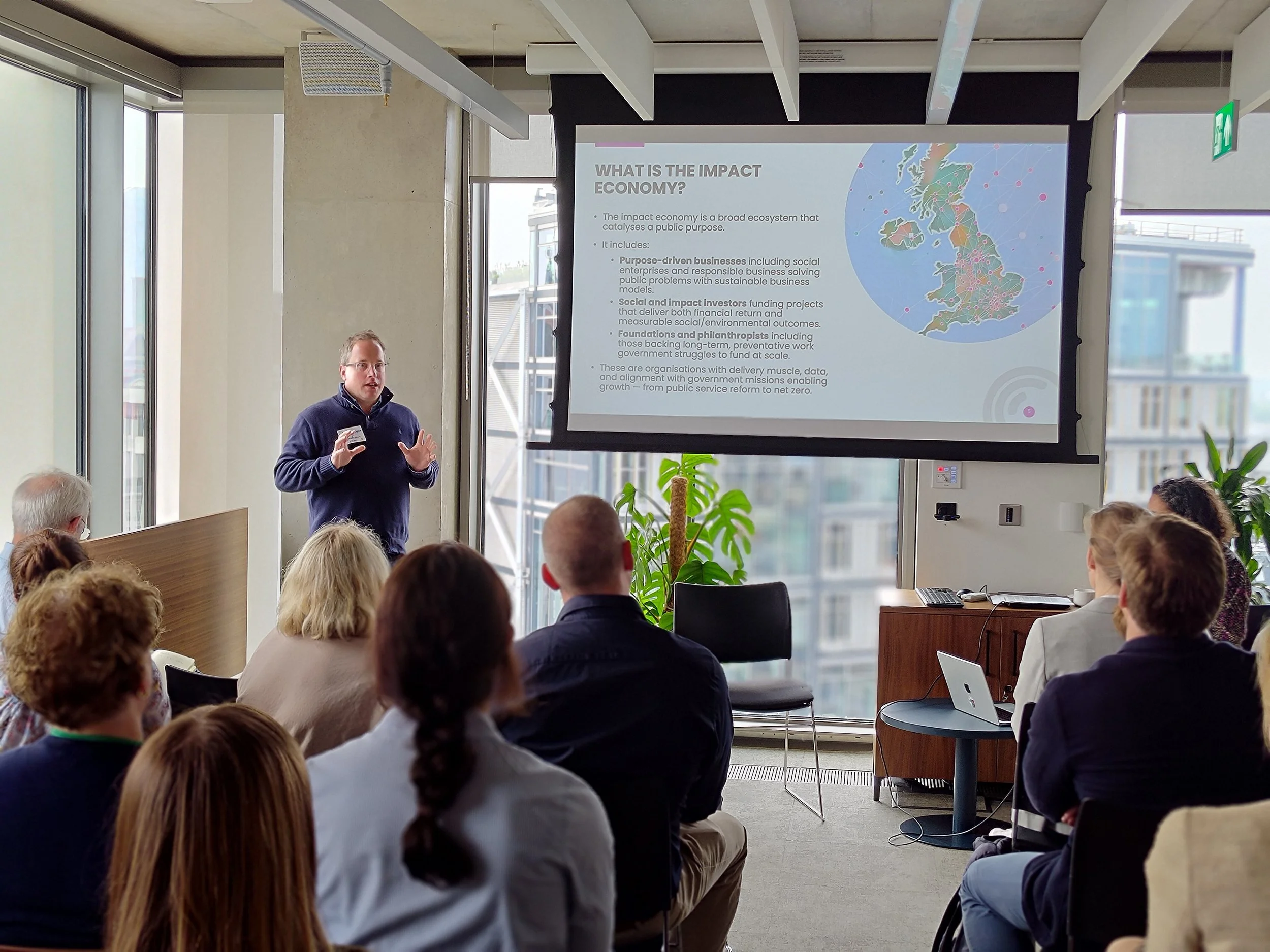 A man giving a presentation on the impact economy in a modern conference room with large windows. The slide on the screen displays a map of Europe and text explaining the impact economy, with a small audience seated and listening.