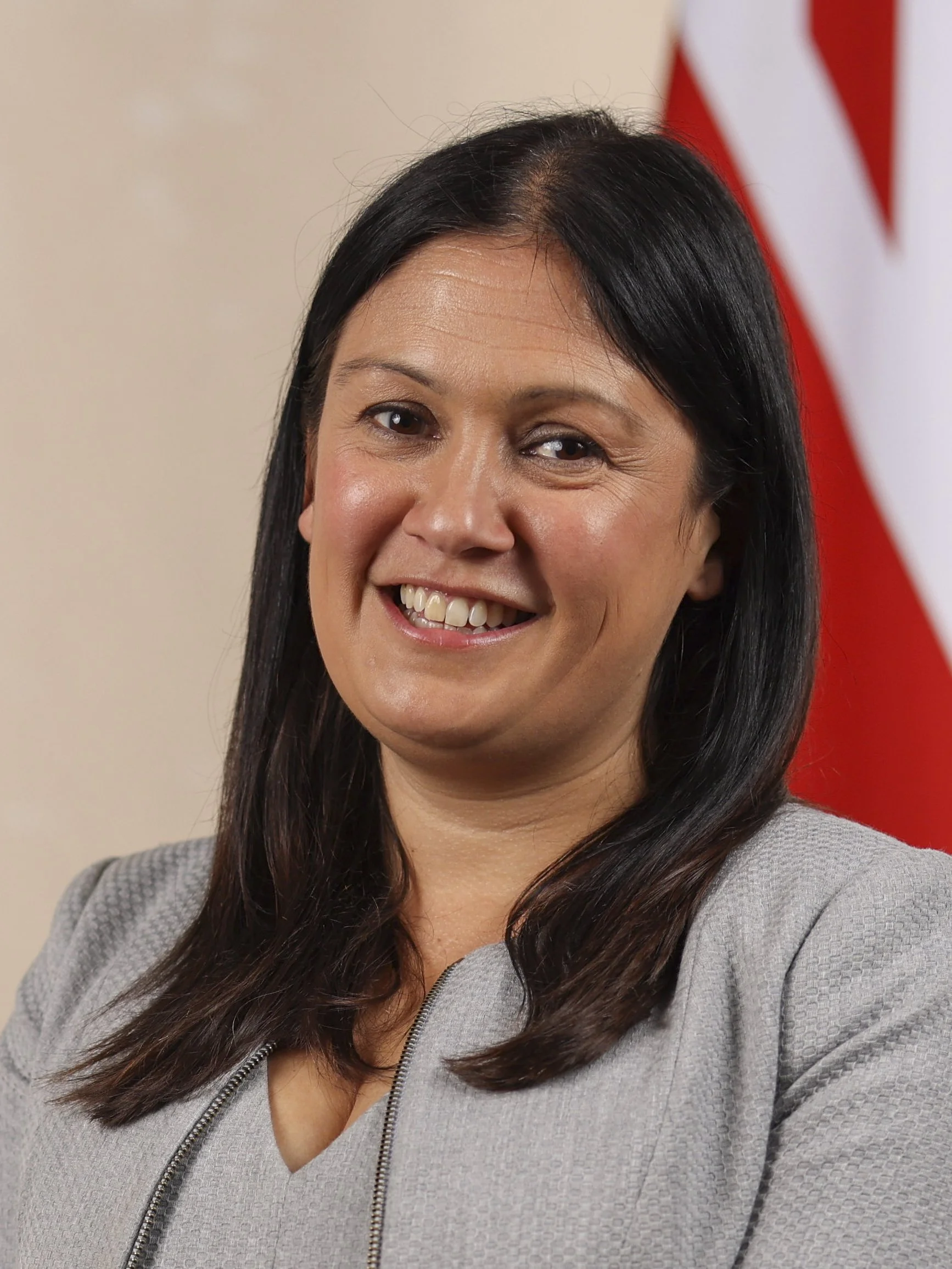 A woman with dark hair wearing a gray blazer, smiling in front of a blurred background with a red and white flag.