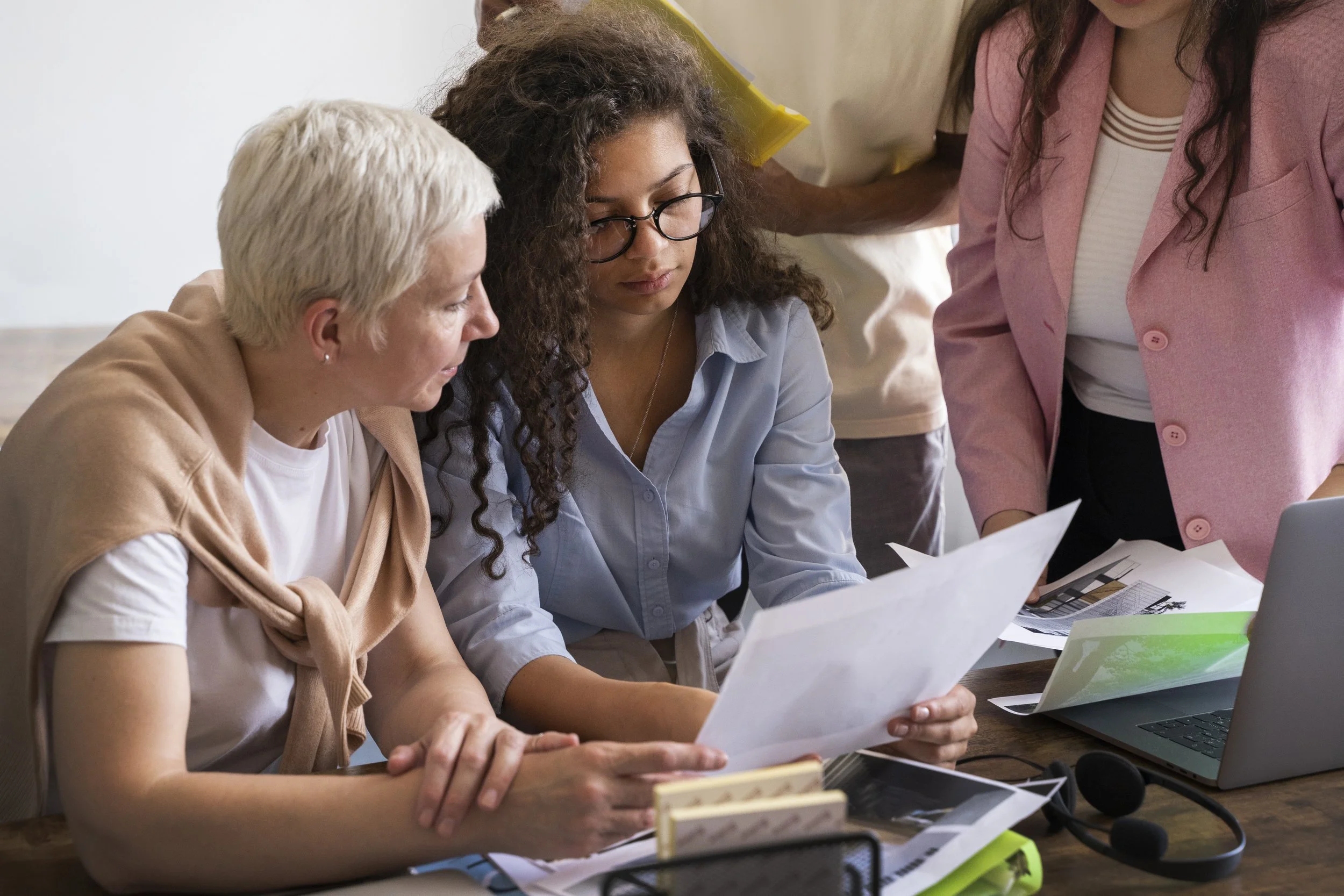 Group of women gathered around a desk reviewing papers and documents, with a laptop and office supplies on the table.