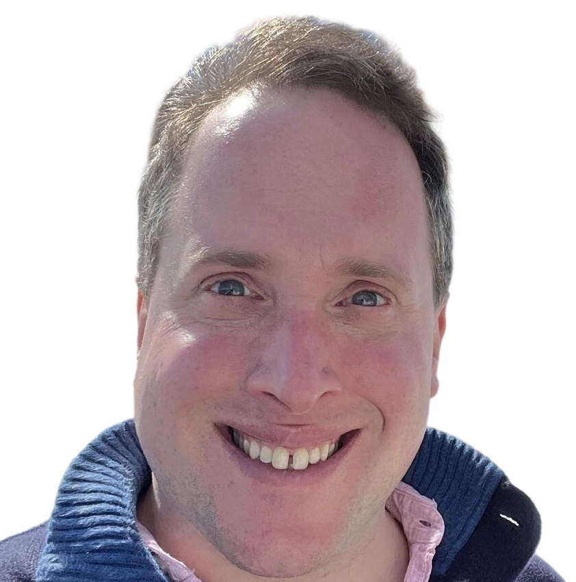 A smiling man with short brown hair, blue eyes, and wearing a navy blue sweater over a light pink shirt, against a plain white background.
