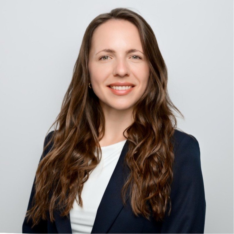 Professional portrait of a smiling woman with long, wavy brown hair, wearing a navy blazer and white top, standing against a light gray background.