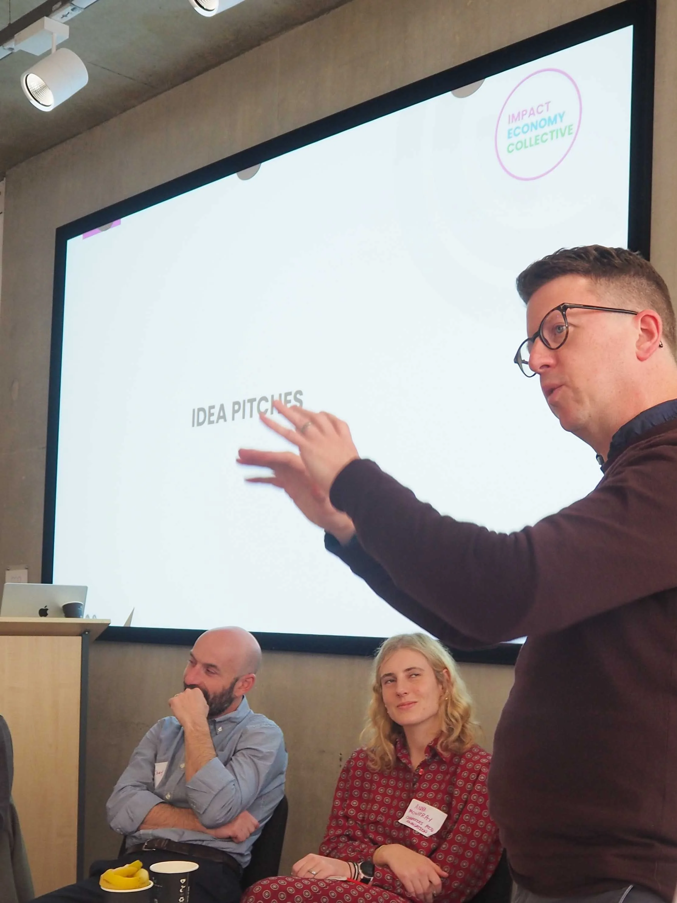A man with glasses and a dark shirt is speaking and gesturing towards a presentation screen saying 'IDEA PITCHES'. Two seated individuals, a man with a beard and a bald head, and a woman with blonde hair, are looking at the speaker. The woman is smiling slightly. The presentation slide shows a logo in the upper right corner with the words 'IMPACT ECONOMY COLLECTIVE' inside a pink circle.