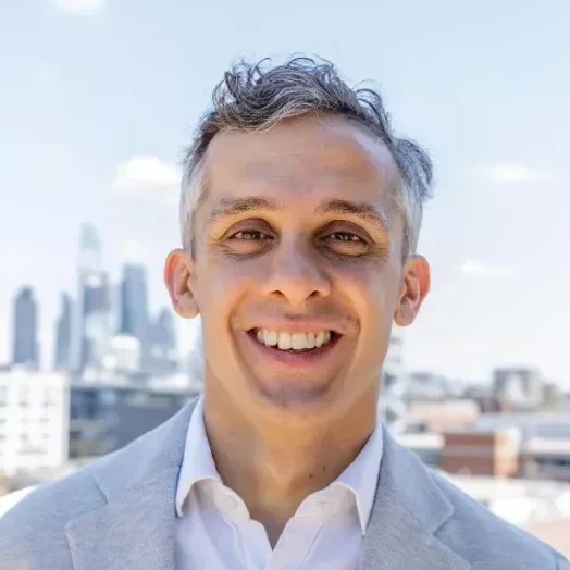 Smiling man in light gray suit jacket and white shirt outdoors in an urban setting with city skyline in the background.