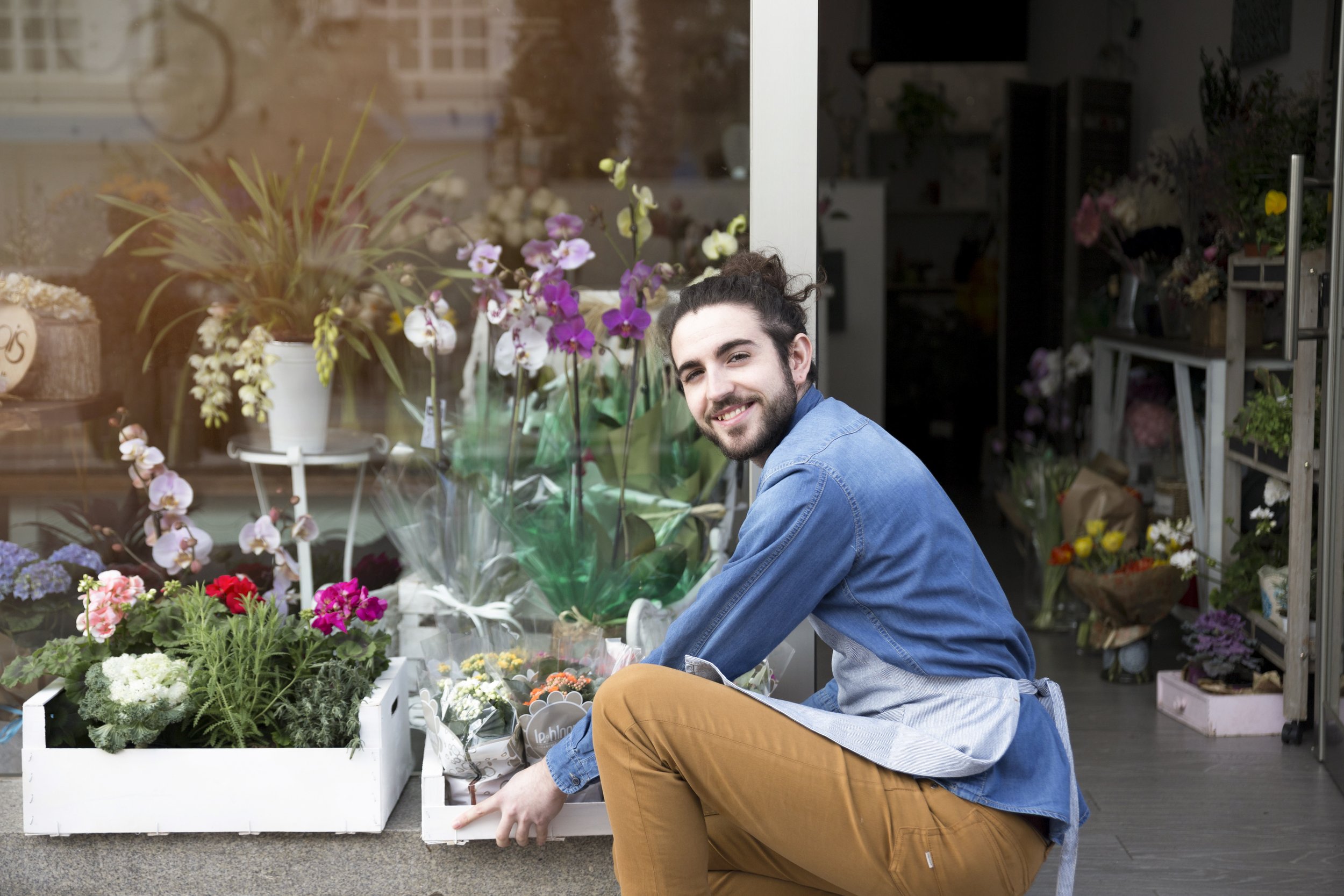A man with dark hair and beard smiling while kneeling outside a flower shop, surrounded by various potted plants and bouquets of colorful flowers.