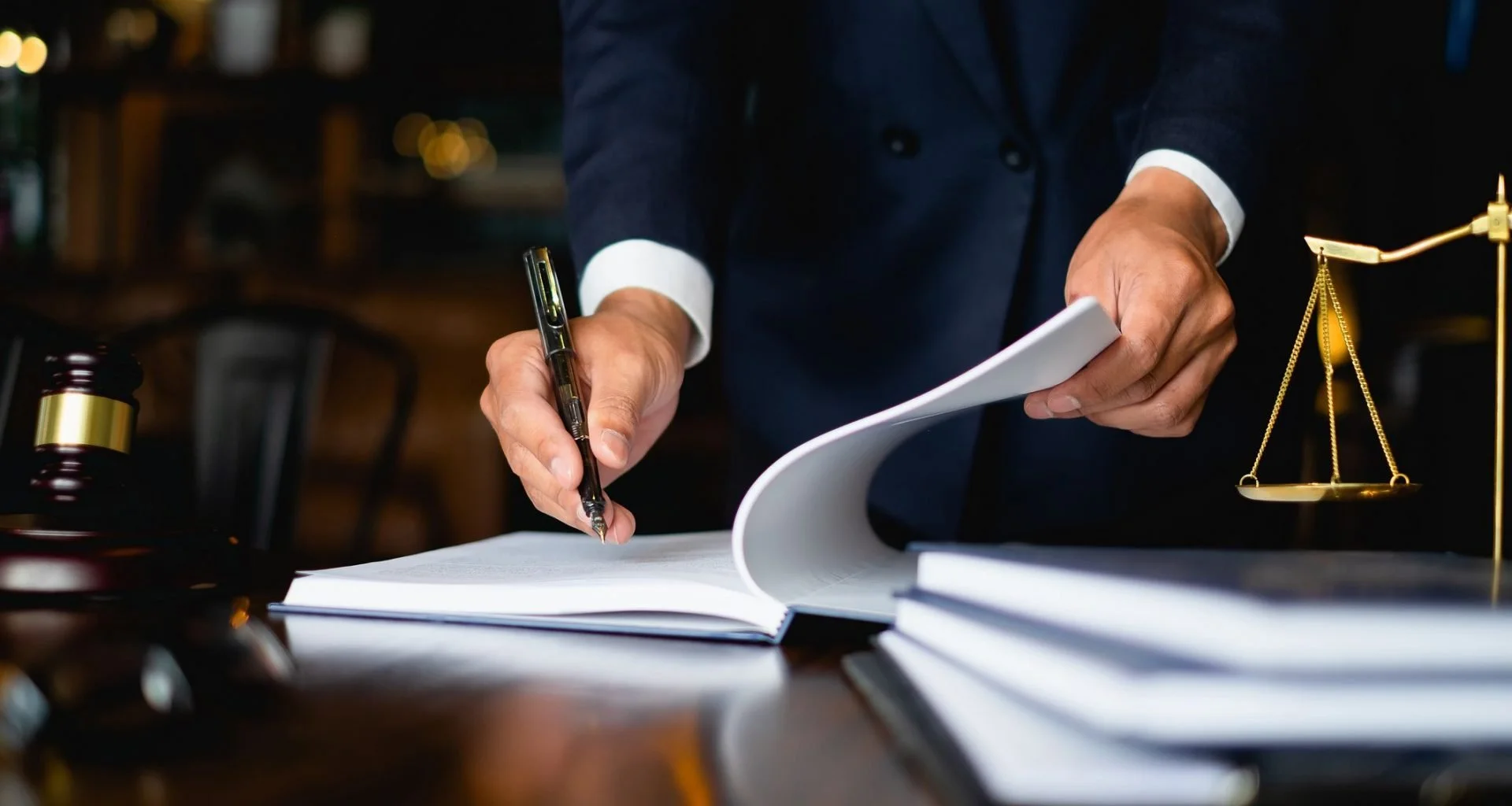A person in a suit writing in a legal pad on a wooden desk with a gavel, a legal scale, and stacks of paper or legal books in the background, indicating a legal or courtroom setting.