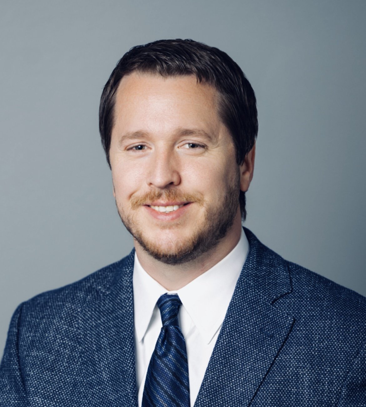 Professional headshot of a man with dark hair, beard, in a suit and tie, smiling, against a plain gray background.