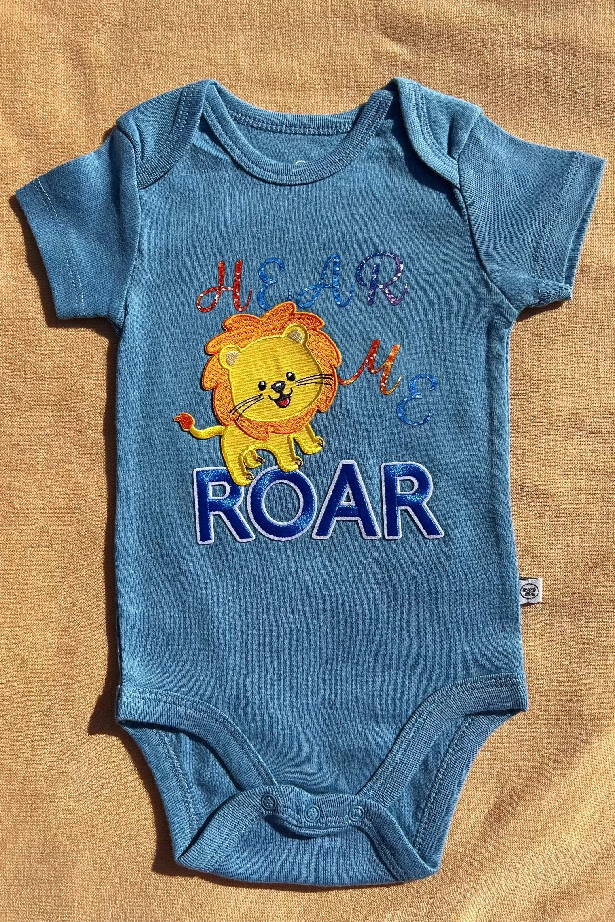 Blue baby onesie with a colorful embroidered design of a smiling lion and the phrase "HEAR ME ROAR".