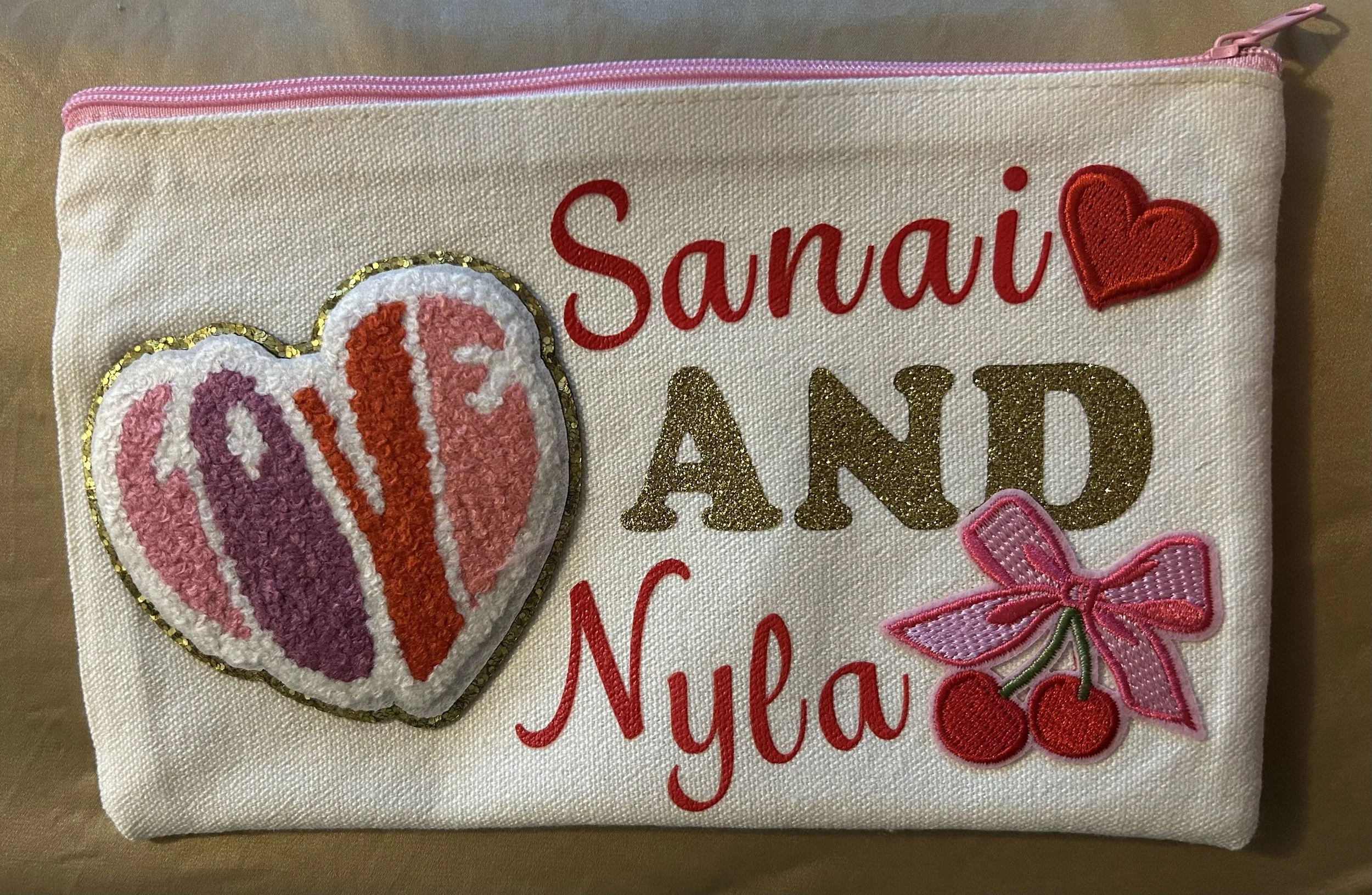 A white fabric pouch decorated with embroidered and patch designs, featuring the text 'Sanai and Nyla' in red and gold glitter, a red heart, a pink bow with cherries, and a heart-shaped applique with the letters 'LOVE' in pink and red.