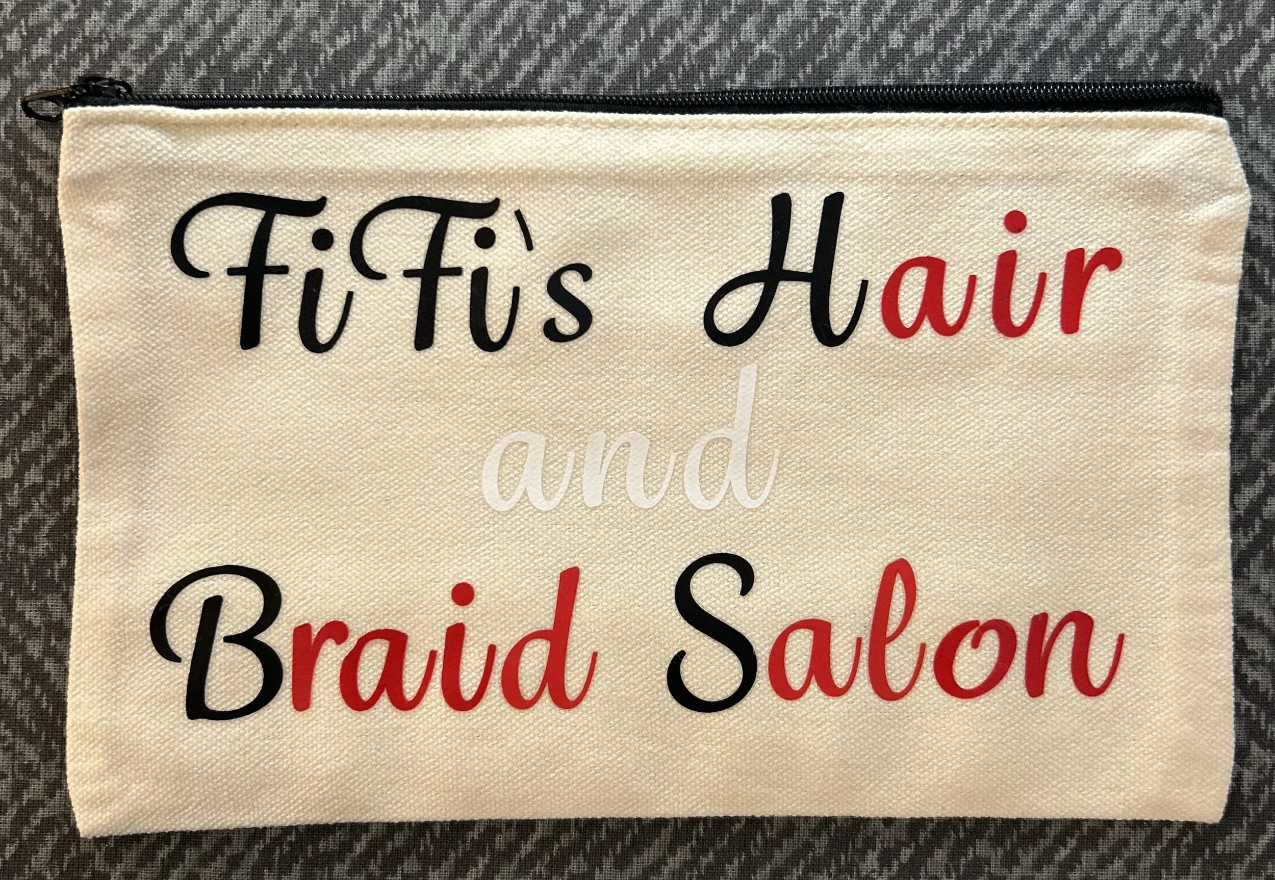 A beige fabric zippered pouch with black, red, and white text that reads 'Fifi's Hair and Braid Salon' on a gray patterned surface.